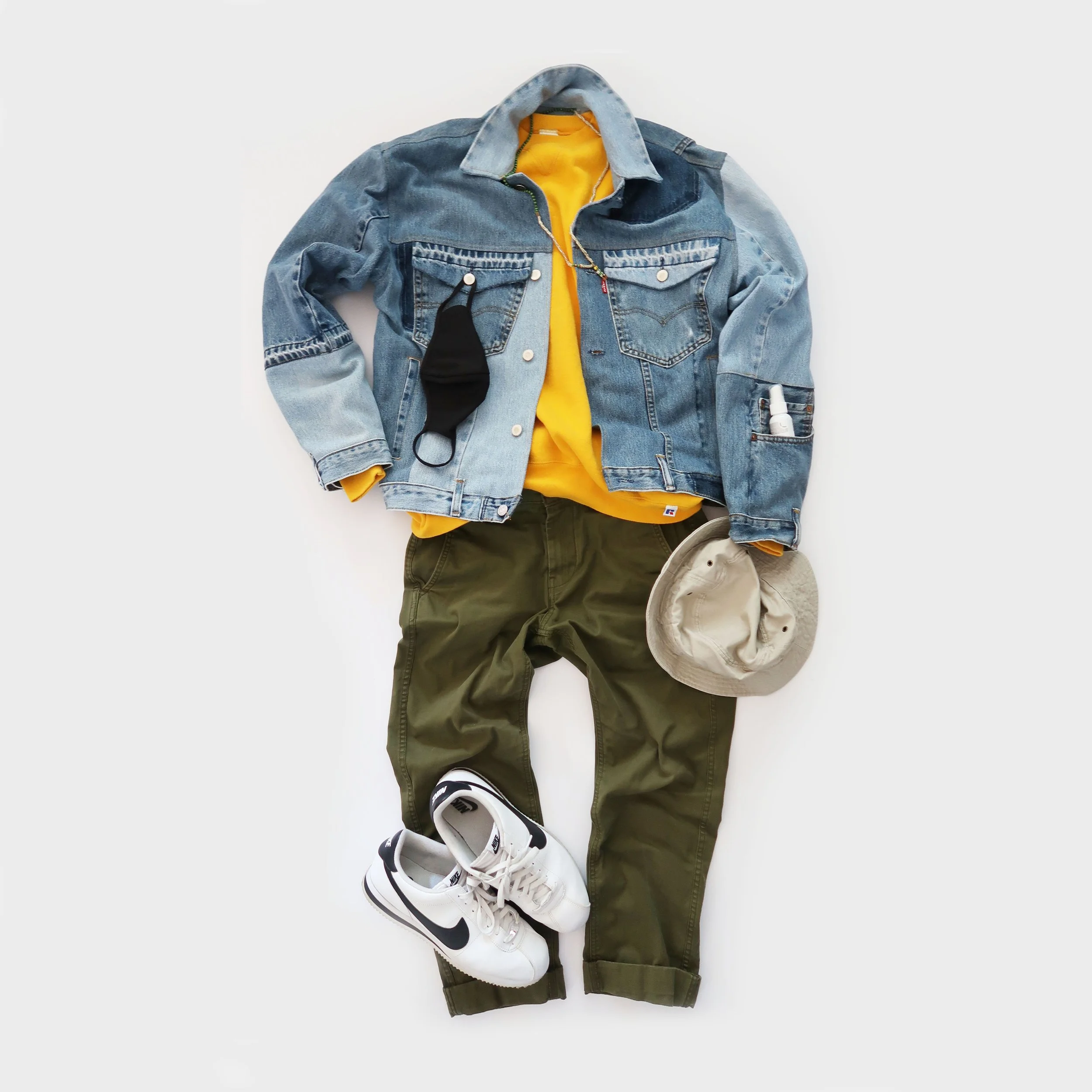 Flat lay of casual clothing and accessories including a denim jacket over a yellow hoodie, green pants, white sneakers, a black face mask, a small spray bottle, and a pair of earbuds, arranged on a white surface.