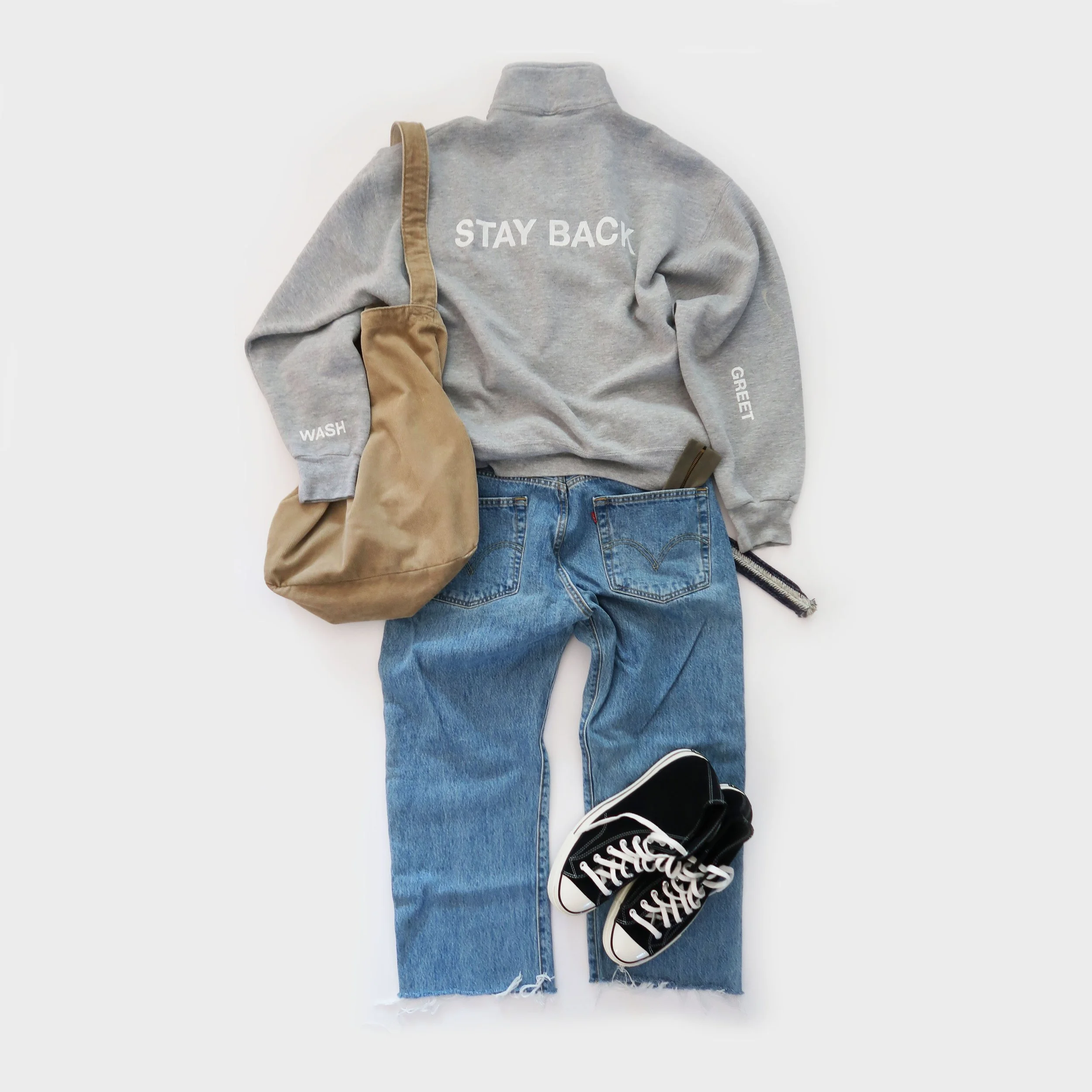 A gray sweatshirt with white text "STAY BACK" on the back, a beige bag, ripped blue jeans, and black sneakers with white laces and soles are laid out on a white background.