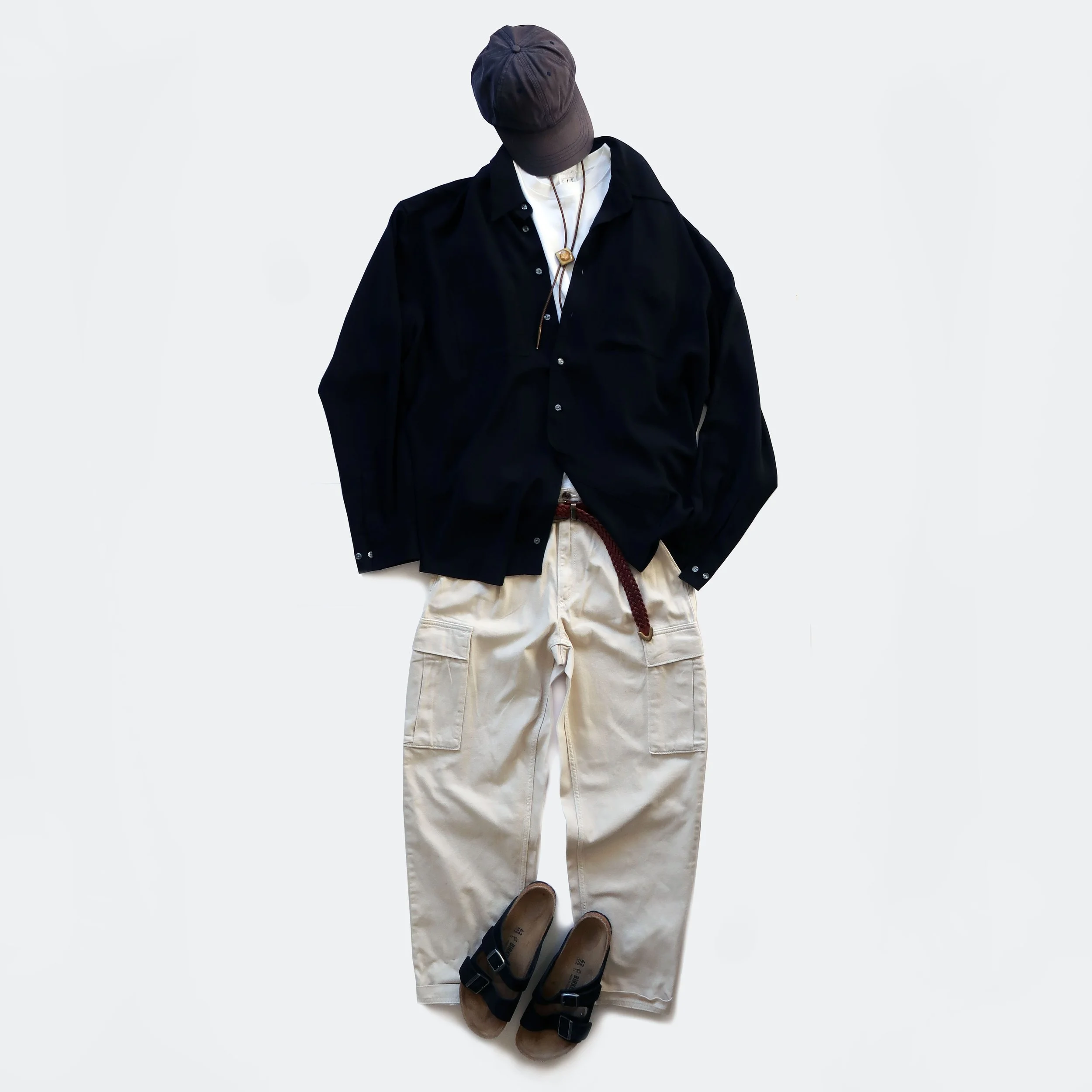 Clothing and footwear arranged on a white background, including a black shirt, beige pants, dark brown sandals, a black hat worn low, and a necklace with a pendant.