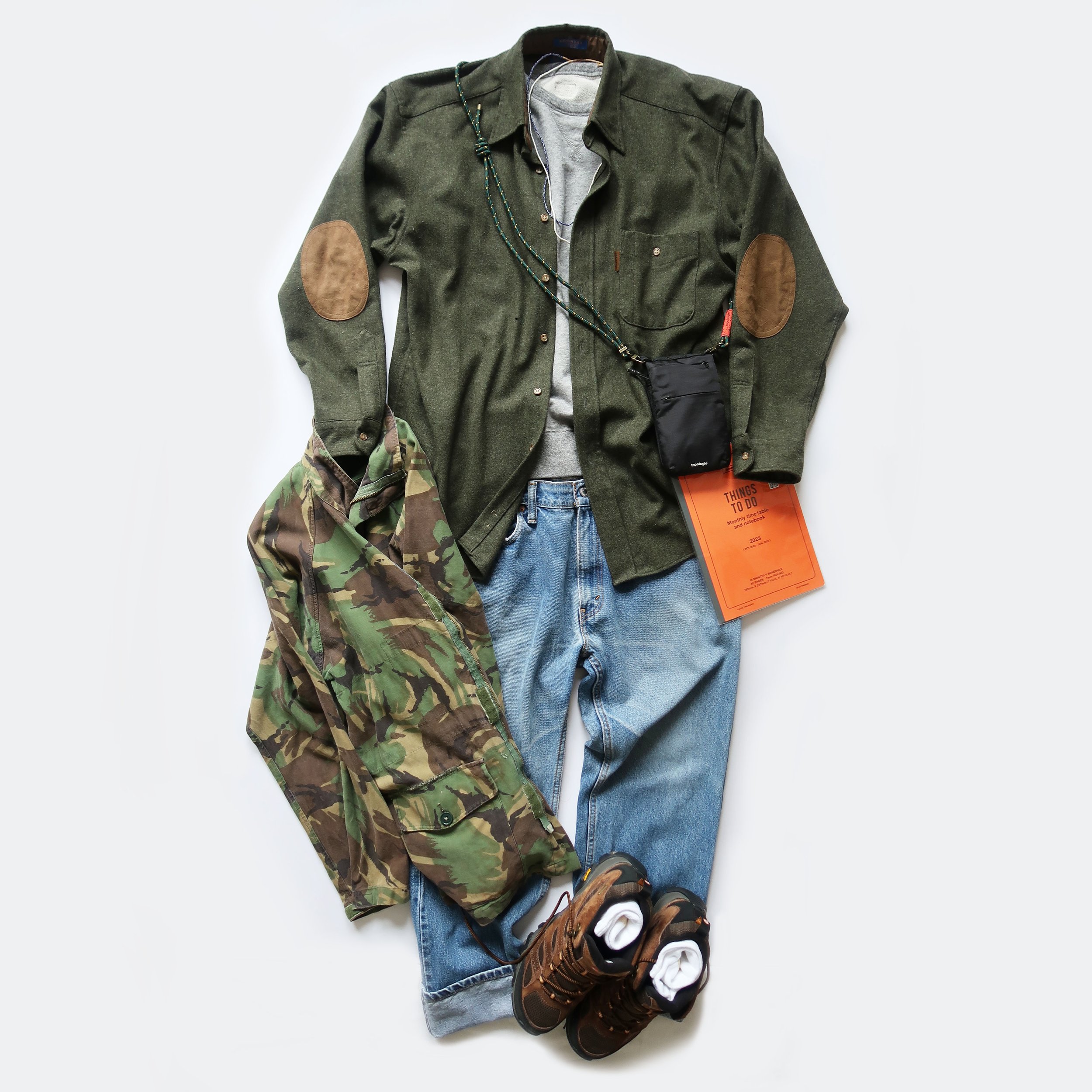 Lay flat display of men's casual and outdoor clothing including a green jacket with leather elbow patches, camouflage pants, a gray sweatshirt, blue jeans, brown hiking boots, white socks, and accessories like a beaded necklace, a small black bag, a 