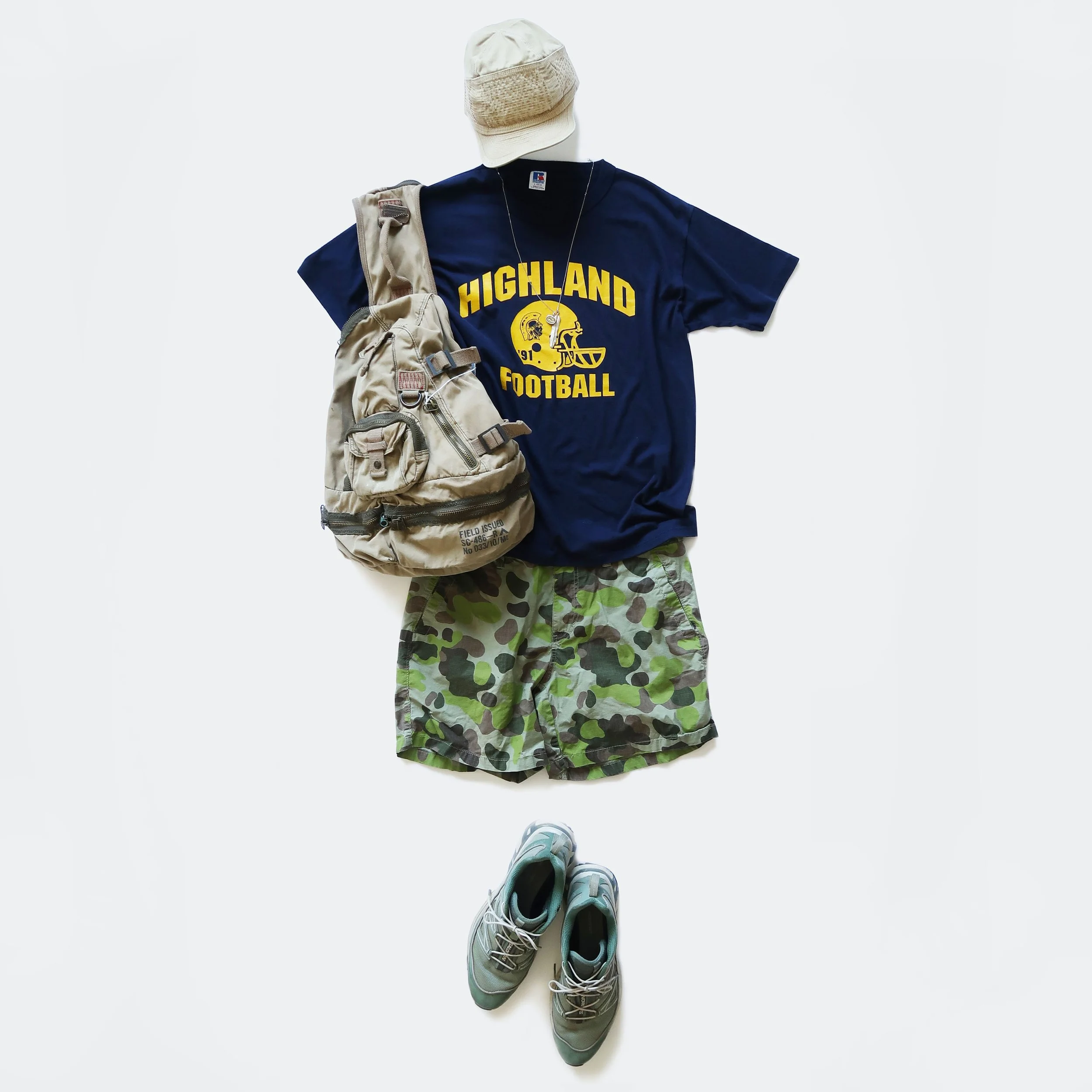Casual athletic outfit with a navy Highland football t-shirt, camouflage shorts, gray running shoes, a beige backpack, a beige cap, and a pair of light blue sneakers seen from a top-down perspective on a white background.
