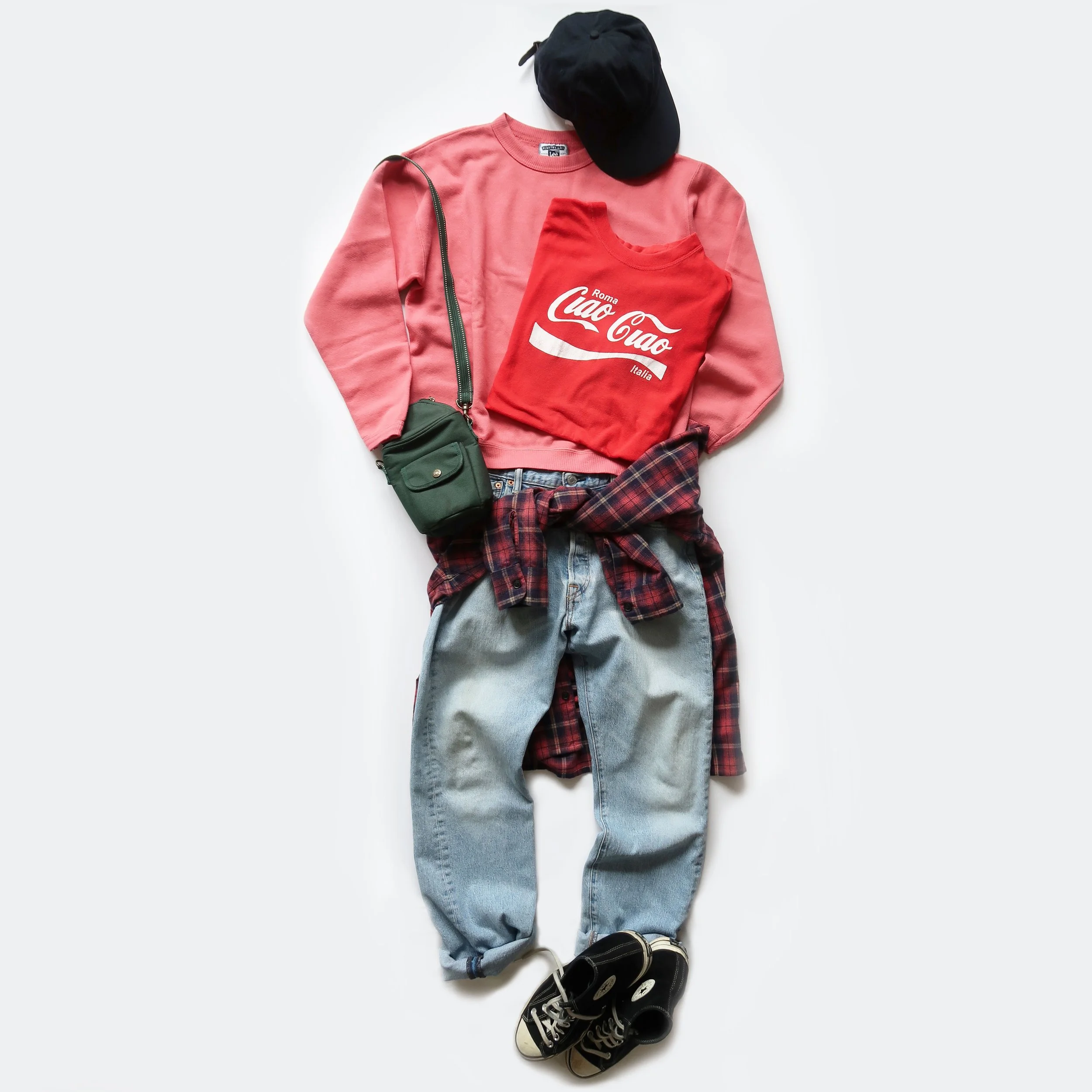 Casual clothing and accessories including a black cap, pink sweatshirt, red graphic T-shirt, plaid shirt, jeans, and black sneakers, arranged on a white surface.