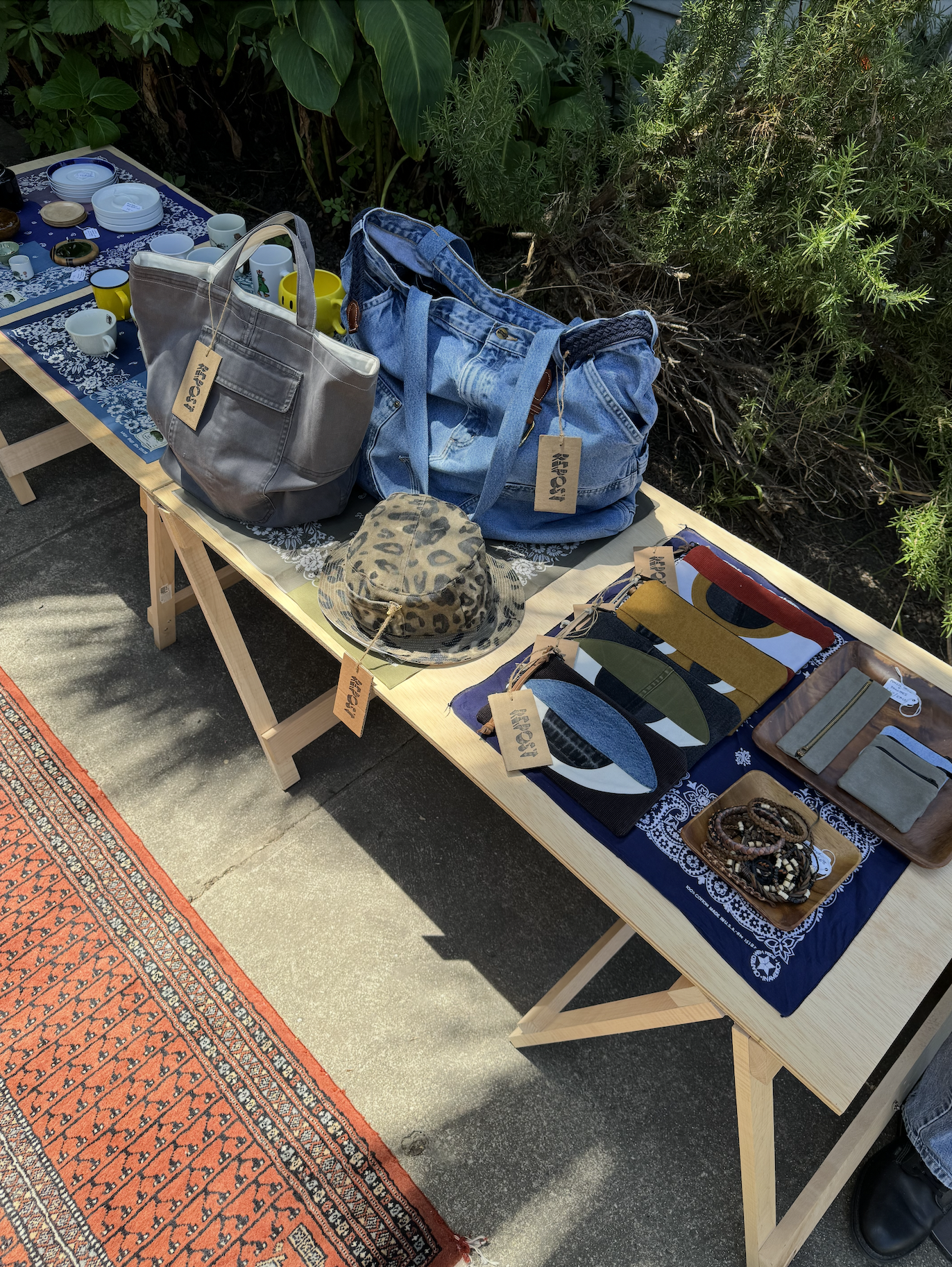 Table with various household items such as dishes, mugs, bags, a hat, and accessories, set outdoors on a sidewalk next to green bushes, with a patterned rug underneath.