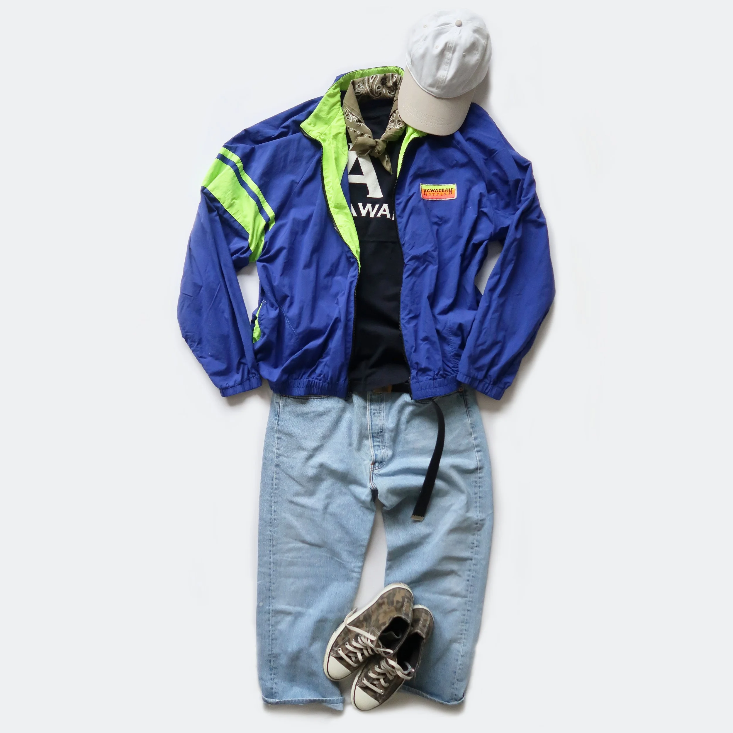 Casual outfit laid out flat on a white surface, including a white cap, black hoodie, blue and green jacket, light blue jeans, and brown sneakers.
