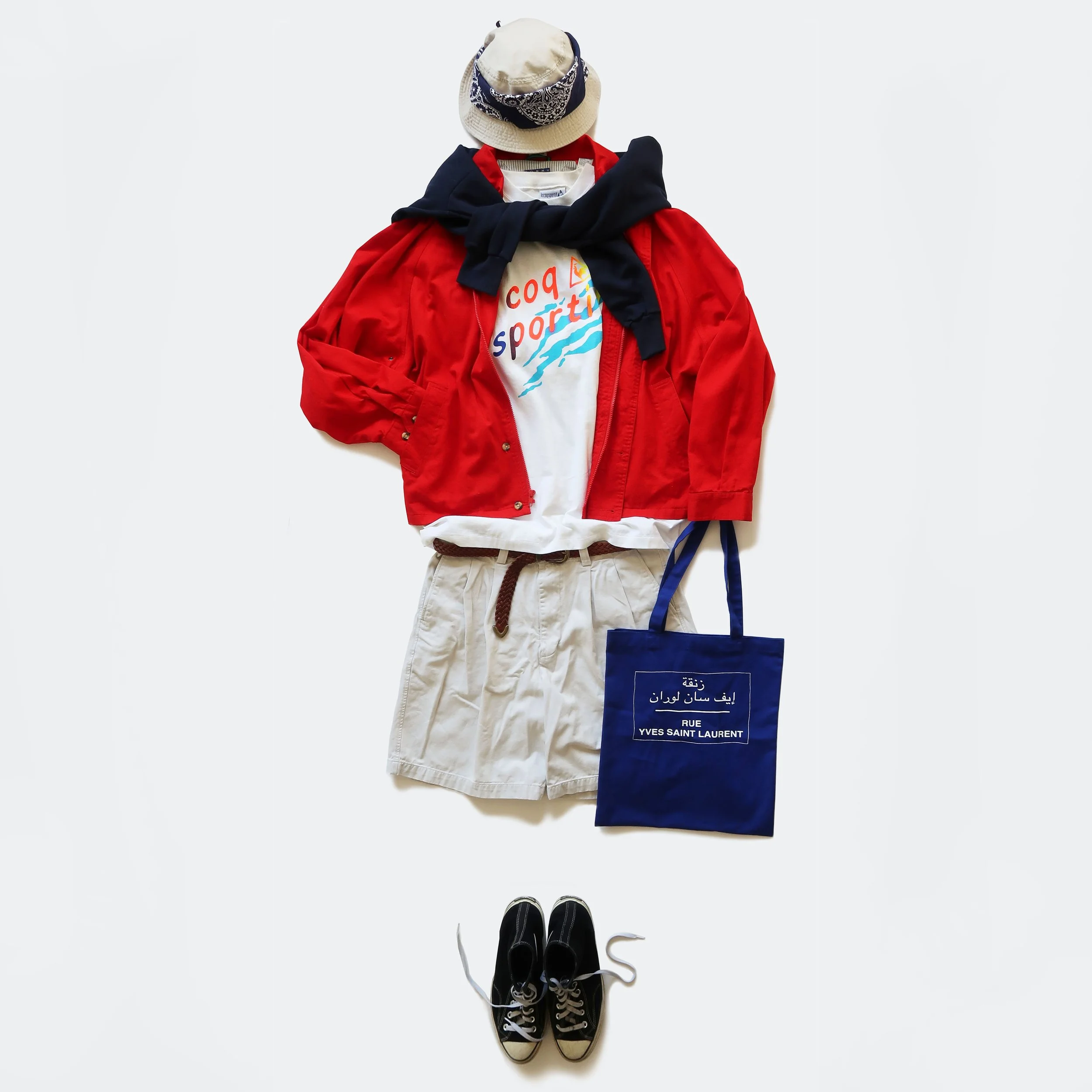 Flat lay of casual clothing and accessories including a beige hat with a bandana, a black sweatshirt, a red jacket, a white t-shirt, beige shorts, a blue bag, and black sneakers, all arranged on a white background.
