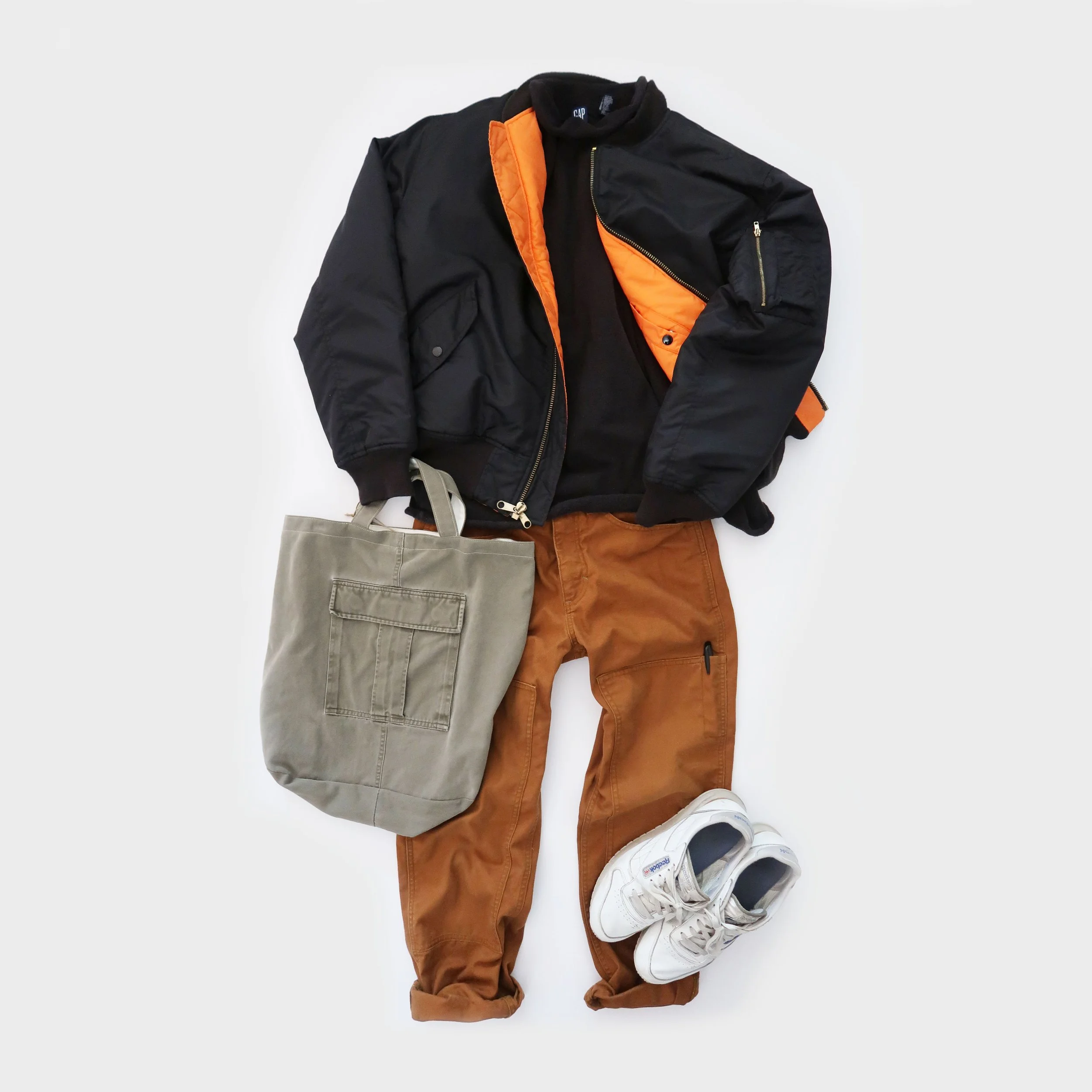 A flat lay of a casual outfit with a black and orange jacket, brown pants, gray tote bag, and white sneakers, arranged on a white surface.