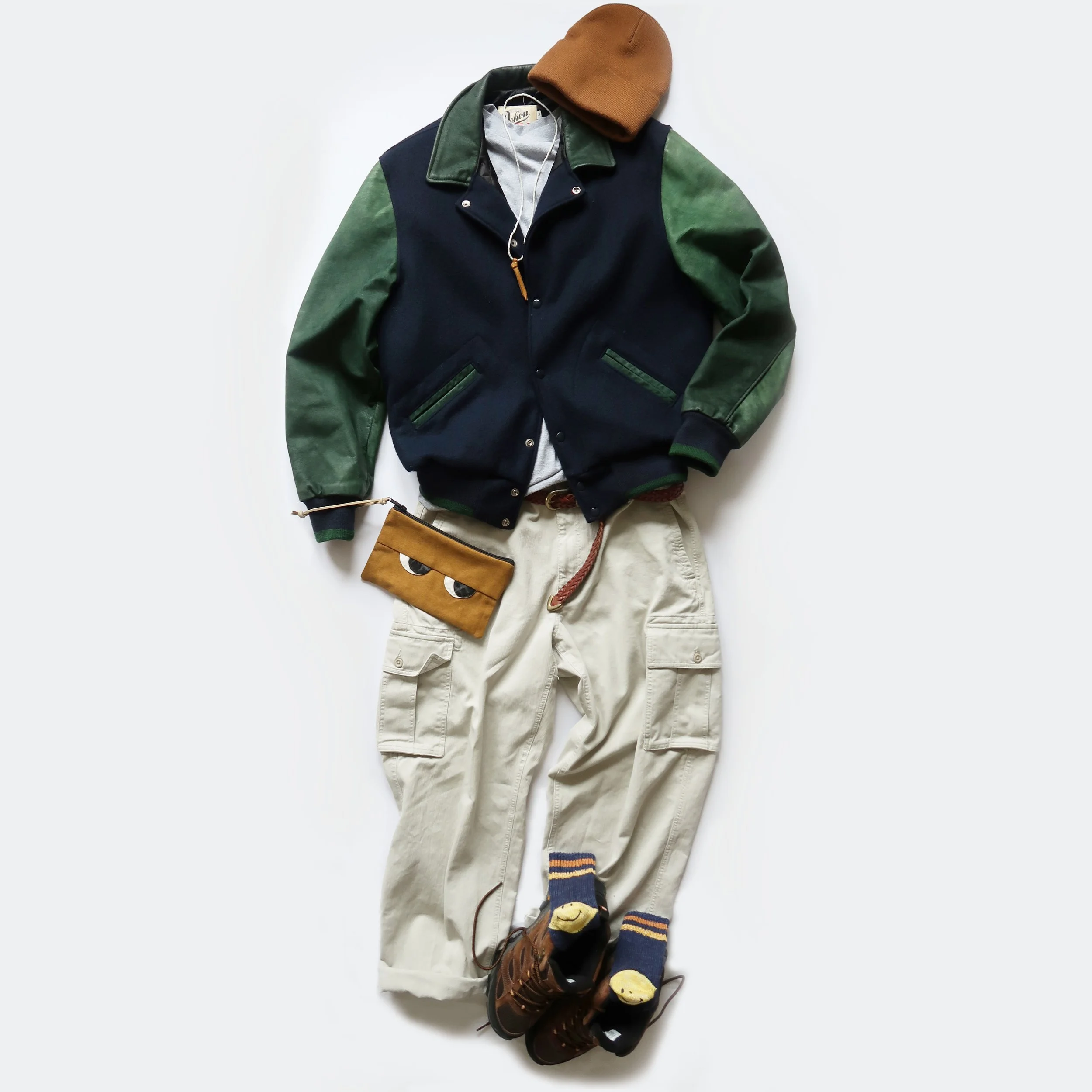 A casual outfit laid out on a white background, including a brown beanie, a navy and green varsity jacket, beige cargo pants, brown hiking boots, and striped socks with smiley faces.