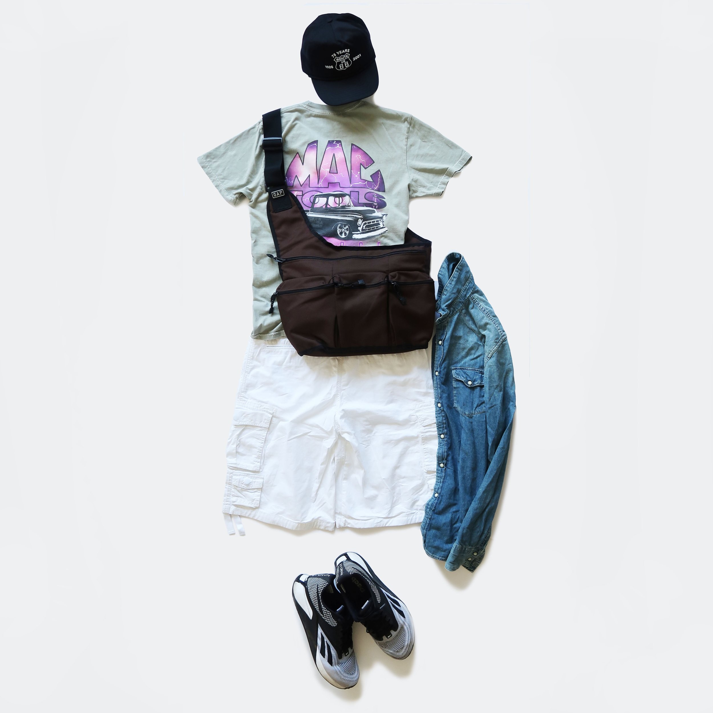 Casual clothing and footwear laid out on a white surface, including a black baseball cap, a graphic T-shirt, denim jeans, athletic shoes, and two bags.
