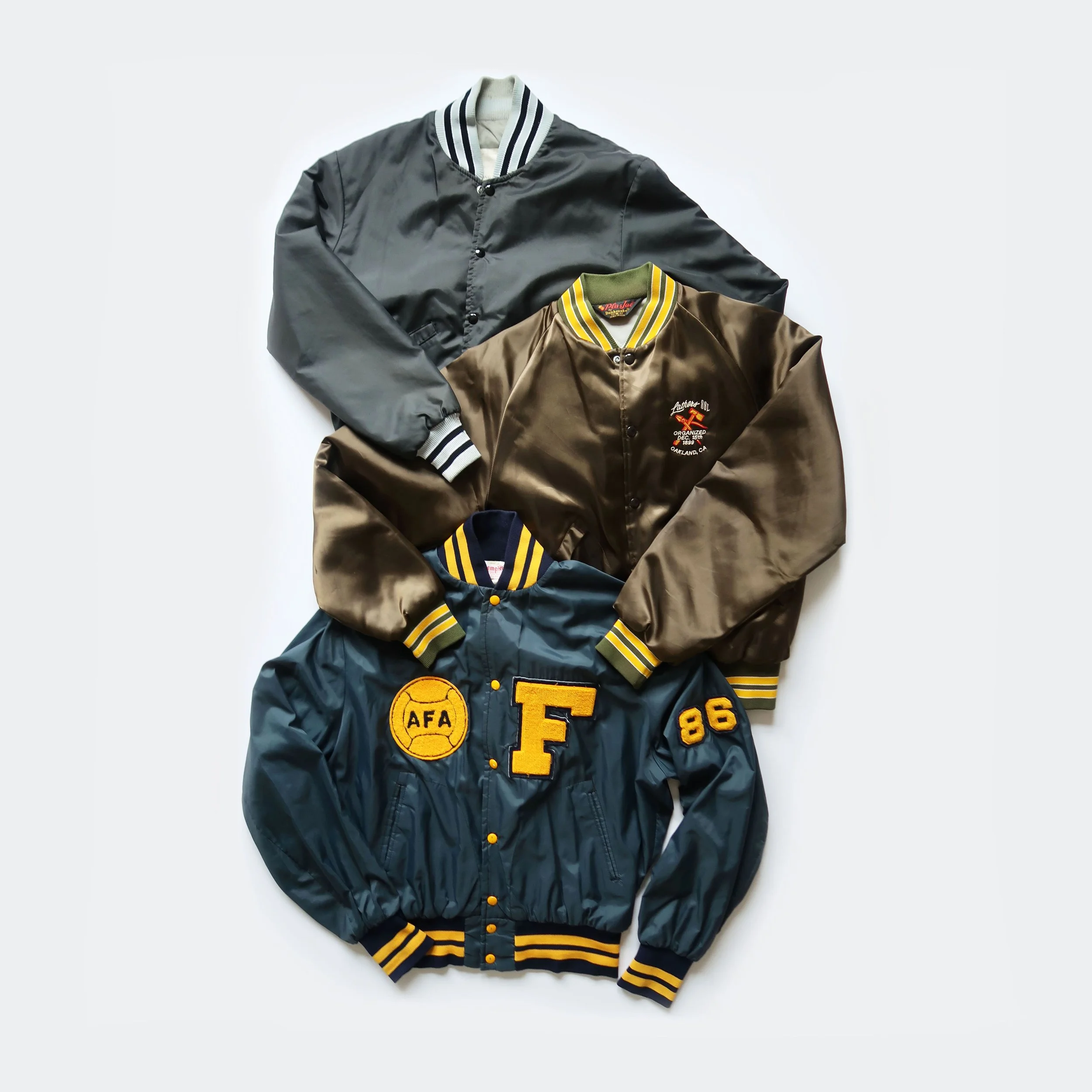 Two varsity jackets, one black and one brown, laid flat against a white background. The jackets feature striped ribbed cuffs and collar, with patches and embroidered designs on the front.