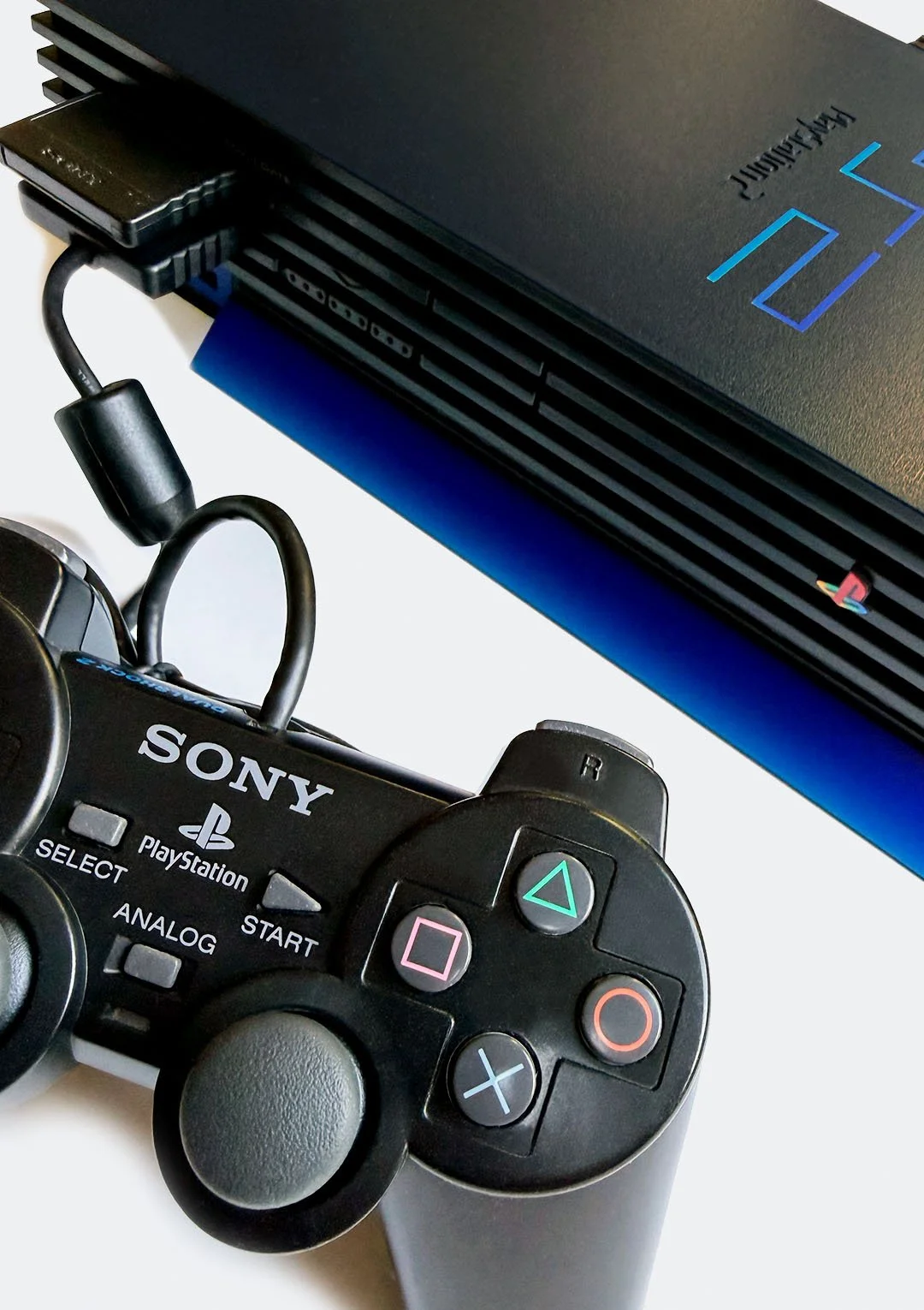 PlayStation 3 console layered on top of a Sony PlayStation controller with black, gray, and multiple colored buttons, on a white surface.