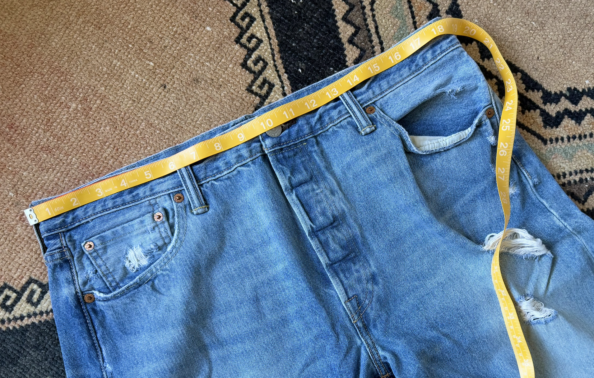 A pair of ripped blue denim jeans lying on a patterned carpet with a yellow measuring tape stretched across the waist.