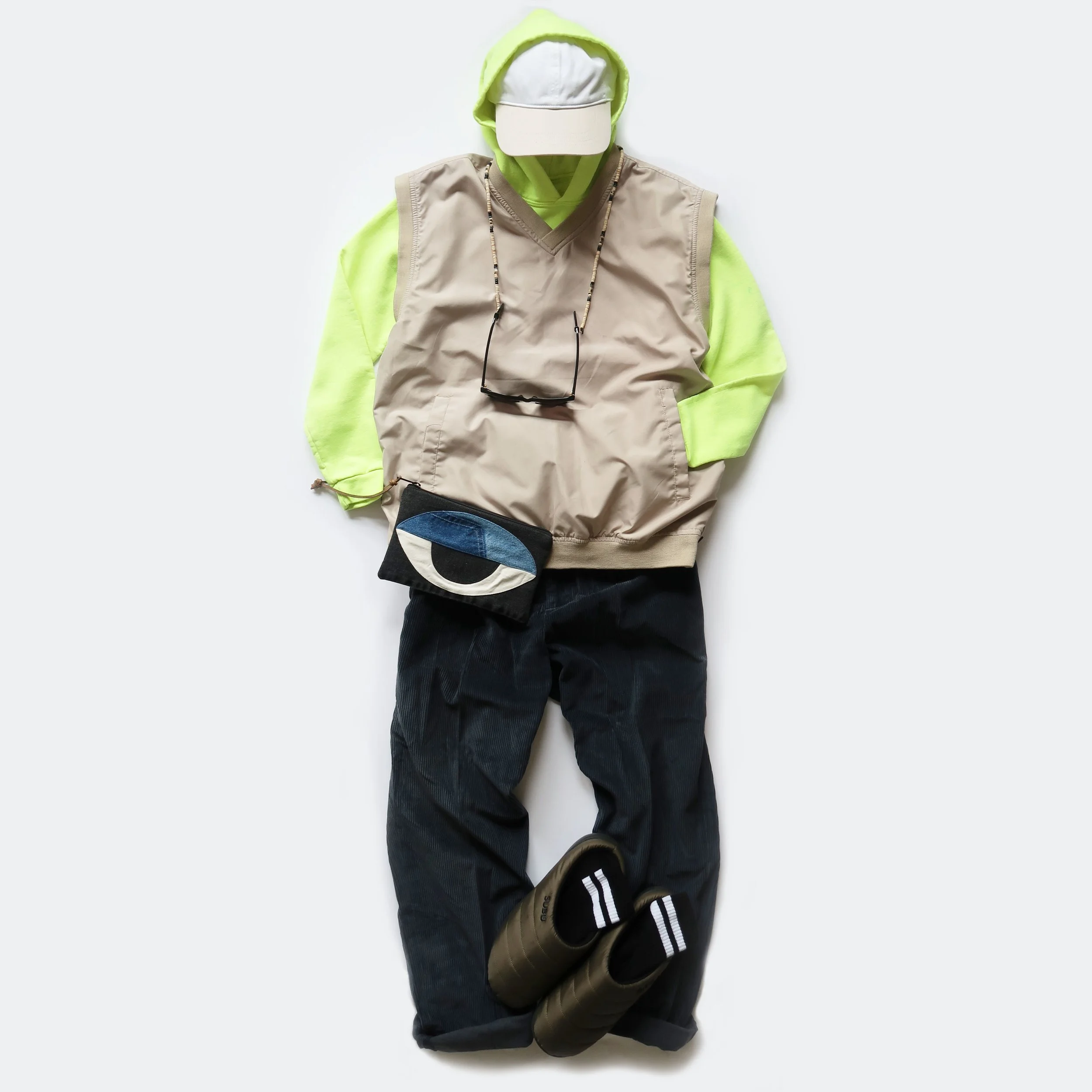 Casual clothing laid out on a white background, including a lime green hoodie, beige vest, black jeans, a pair of black and white sneakers, a beaded necklace, a black crossbody bag, and a beige hat.