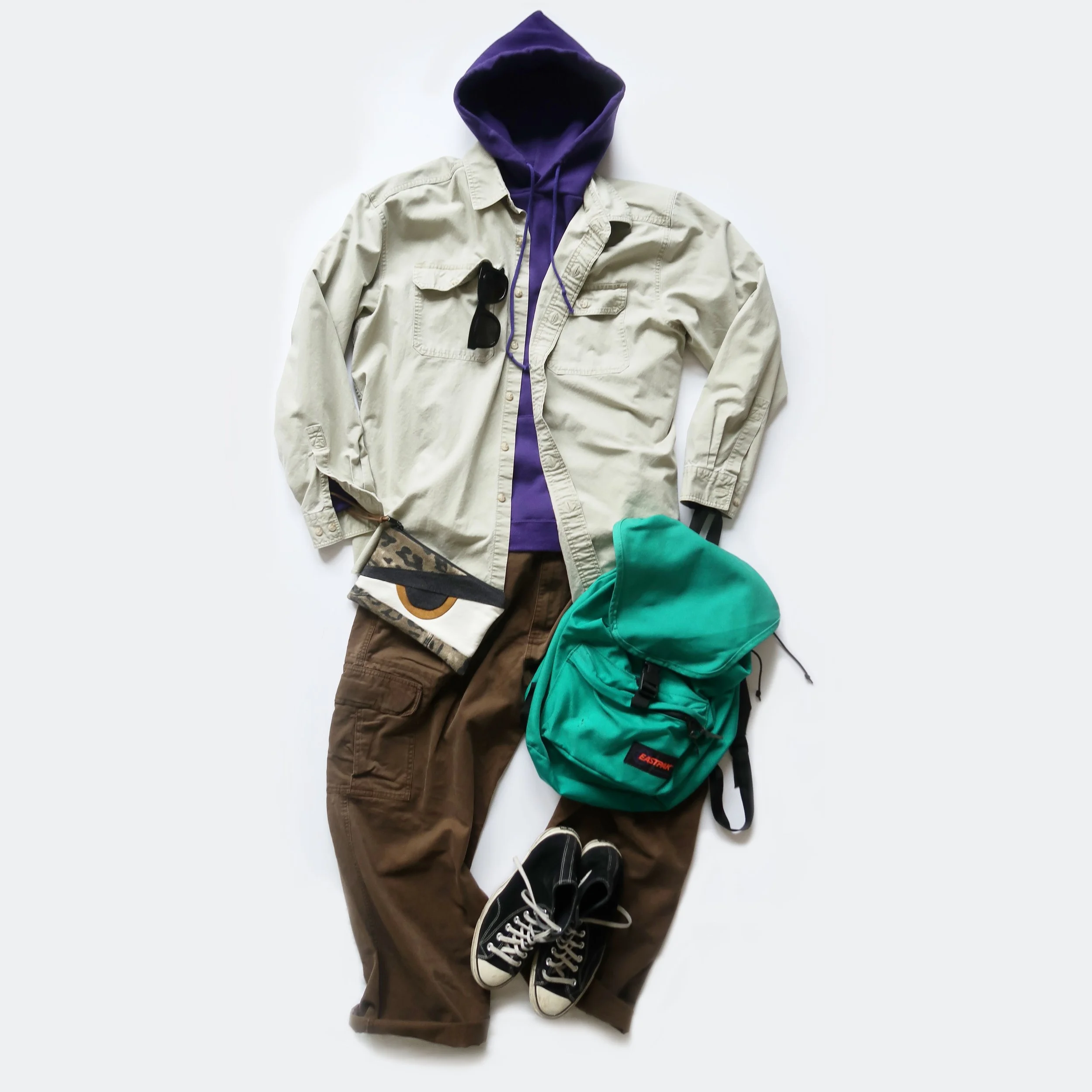 Casual clothing and accessories laid out on a white background, including a purple hoodie under a beige jacket, sunglasses, a patterned cap, green and black backpacks, brown pants, and black Converse sneakers.