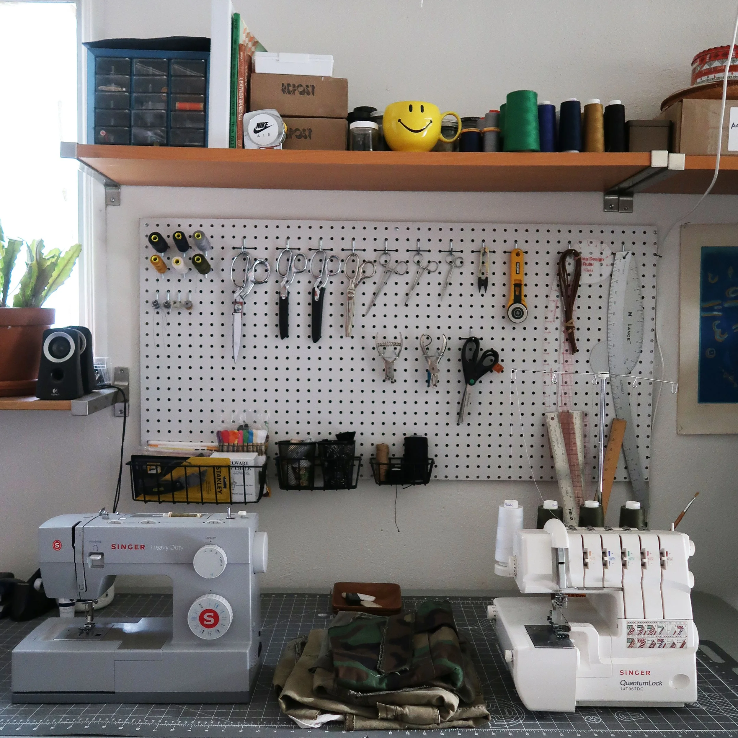 A workspace consisting of two sewing machines, a sewing area with various tools and supplies, and a pegboard with scissors, rulers, and other sewing accessories. The upper shelf holds boxes, thread spools, and a yellow mug with a smiley face, along w