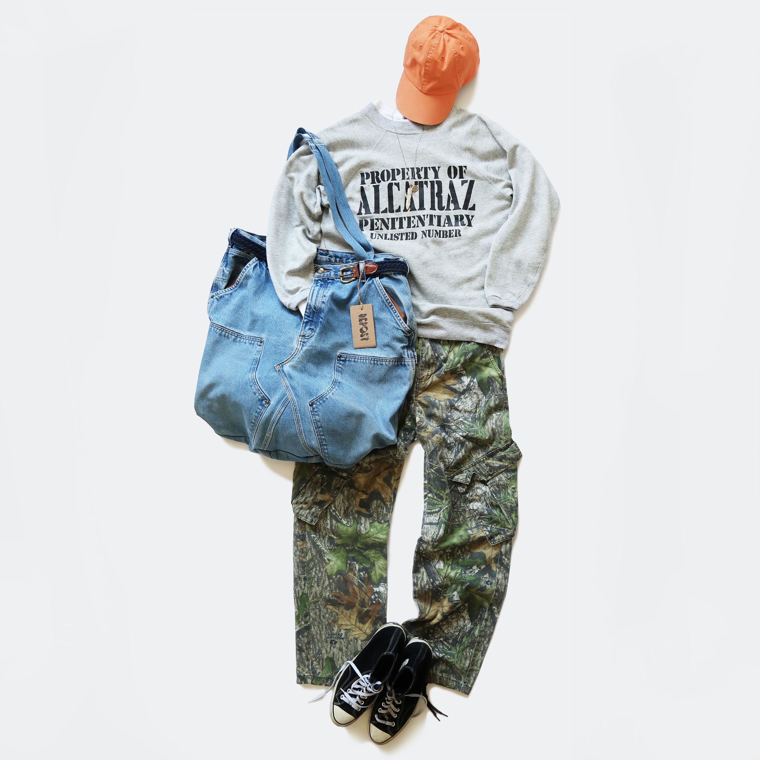 Casual outfit including a gray sweatshirt with text, camouflage pants, black sneakers with white soles, an orange cap, and a denim jacket laid out on a white background.