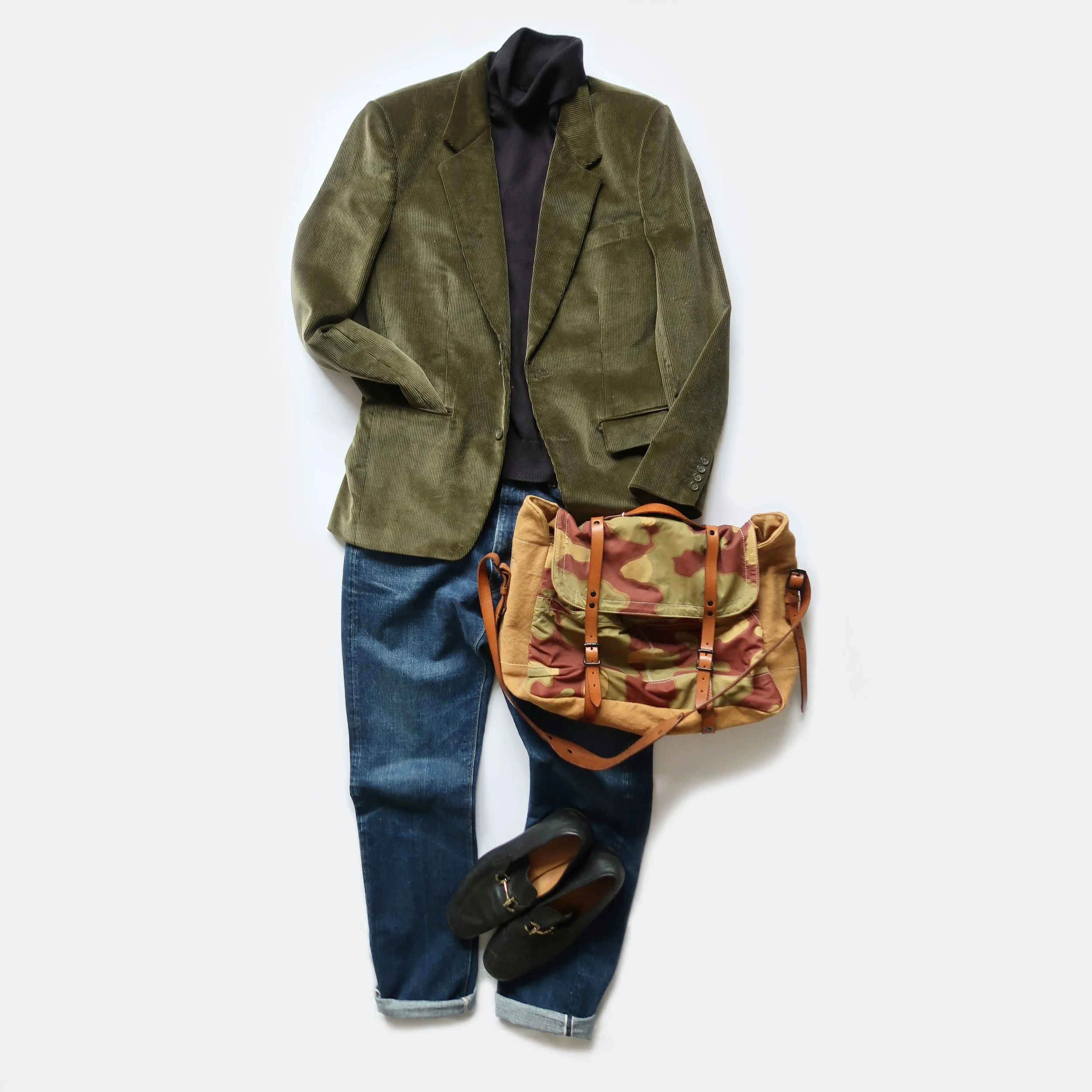A green corduroy blazer layered over a black shirt, paired with blue jeans, black boots, and a camouflage backpack.