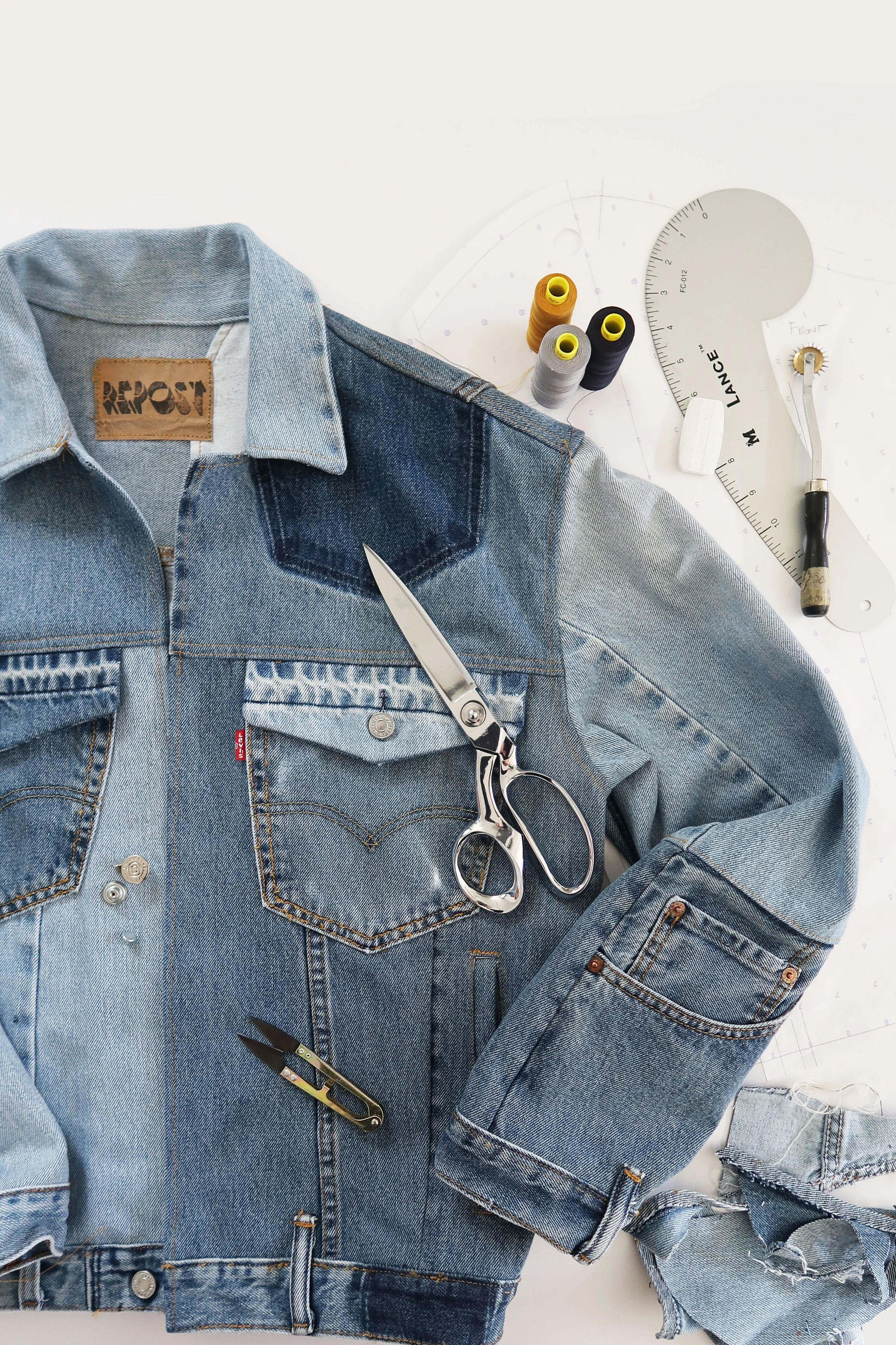 A partially assembled denim jacket with sewing tools, threads, and patterns on a white table.