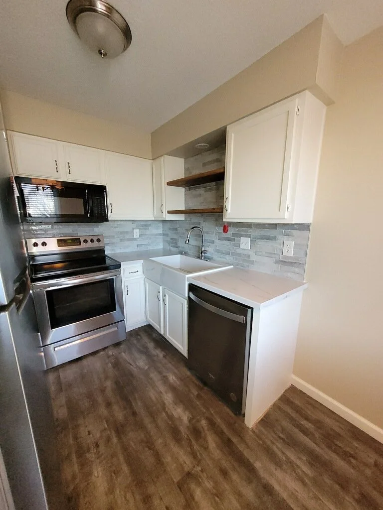 Compact kitchen with stainless steel stove and microwave, white upper and lower cabinets, a white farmhouse sink, a small refrigerator, and open wooden shelves with a grey brick backsplash.