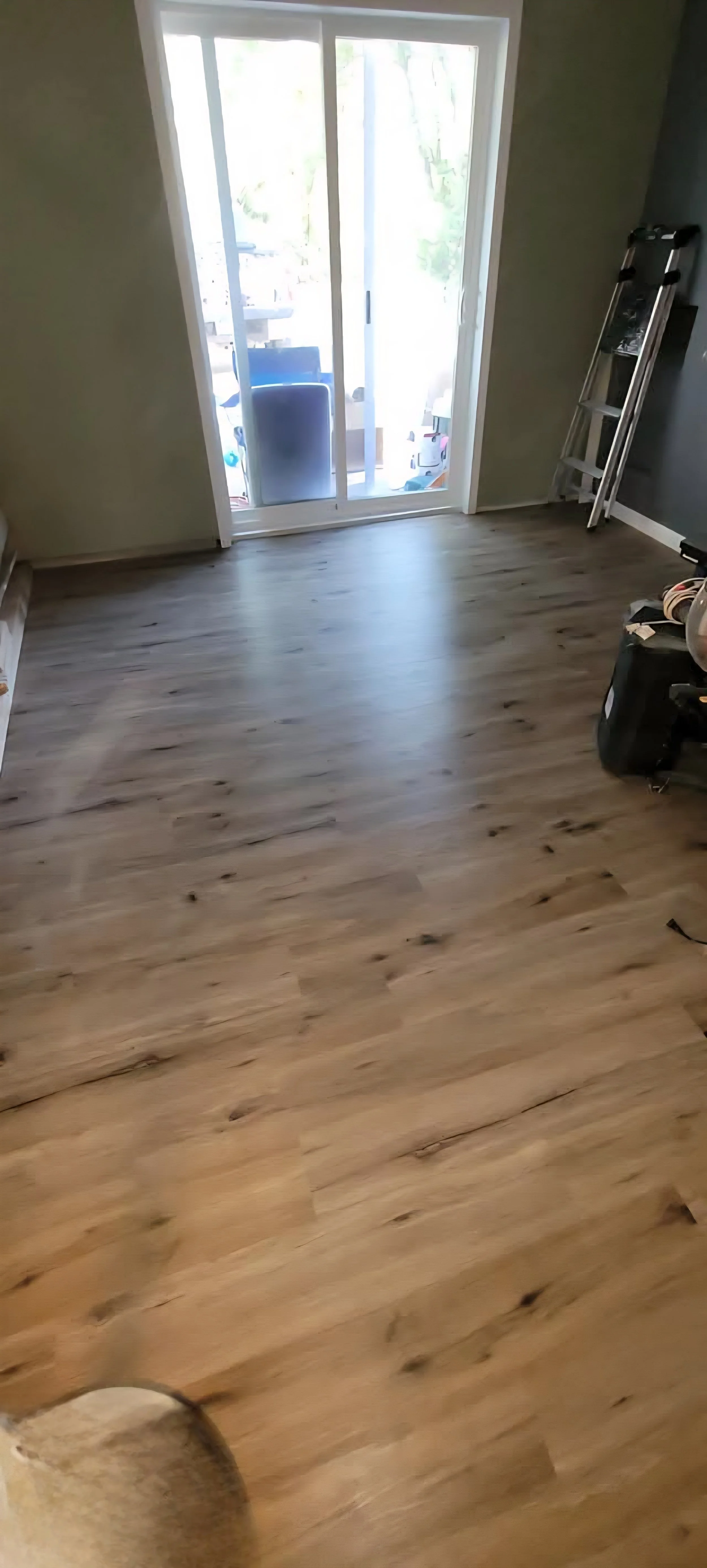 Room with sliding glass door leading to an outdoor patio, light-colored wooden flooring, ladder, and some tools or equipment.