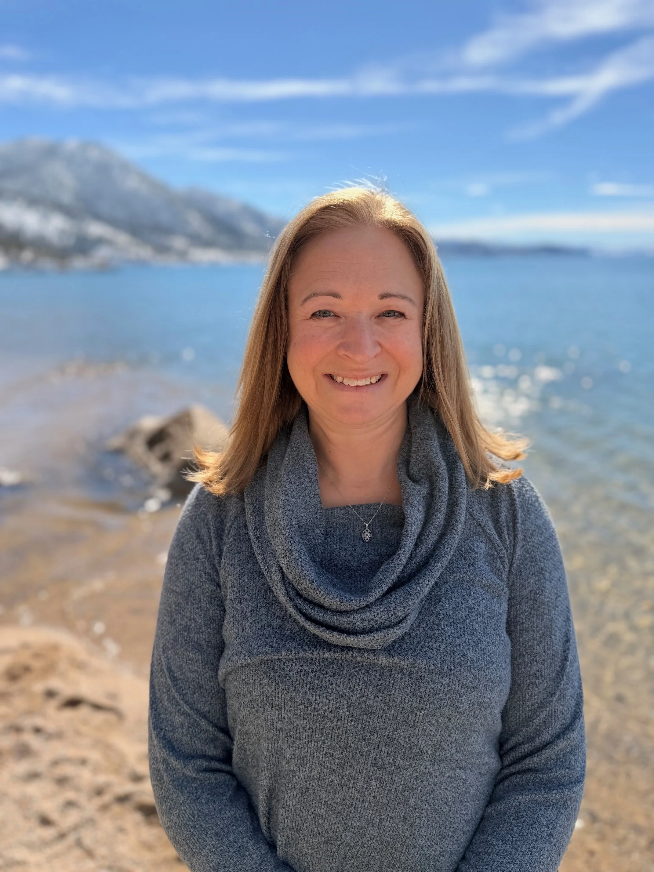 A woman with blonde hair smiling at the camera on a beach, with mountains and a blue sky in the background.