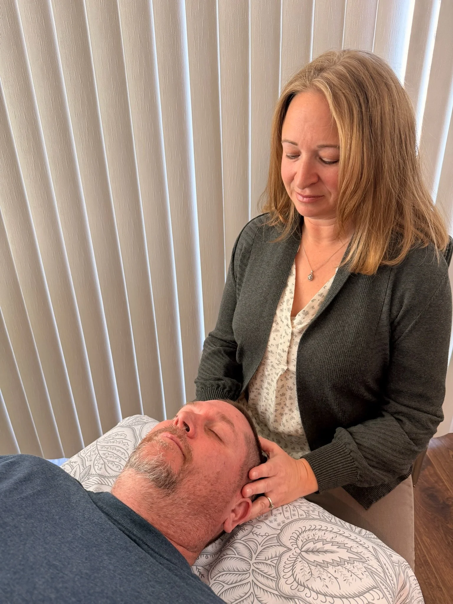 A woman touching a man's head and providing a CranioSacral therapy Treatment while the man is lying on a massage table with closed eyes.
