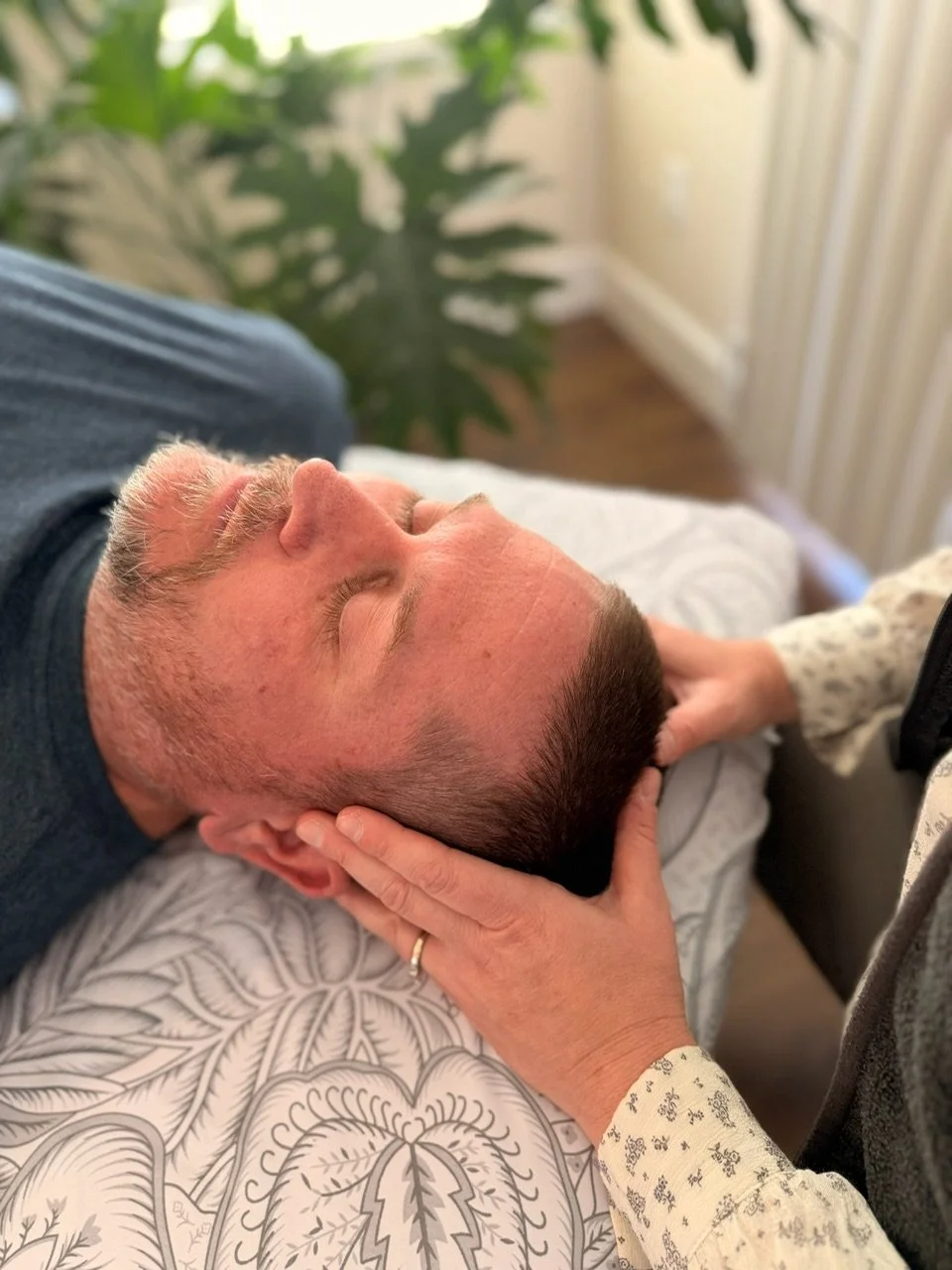 Man lying on a massage table while getting a CranioSacral Therapy treatment from a woman, with a large green plant and curtains in the background.