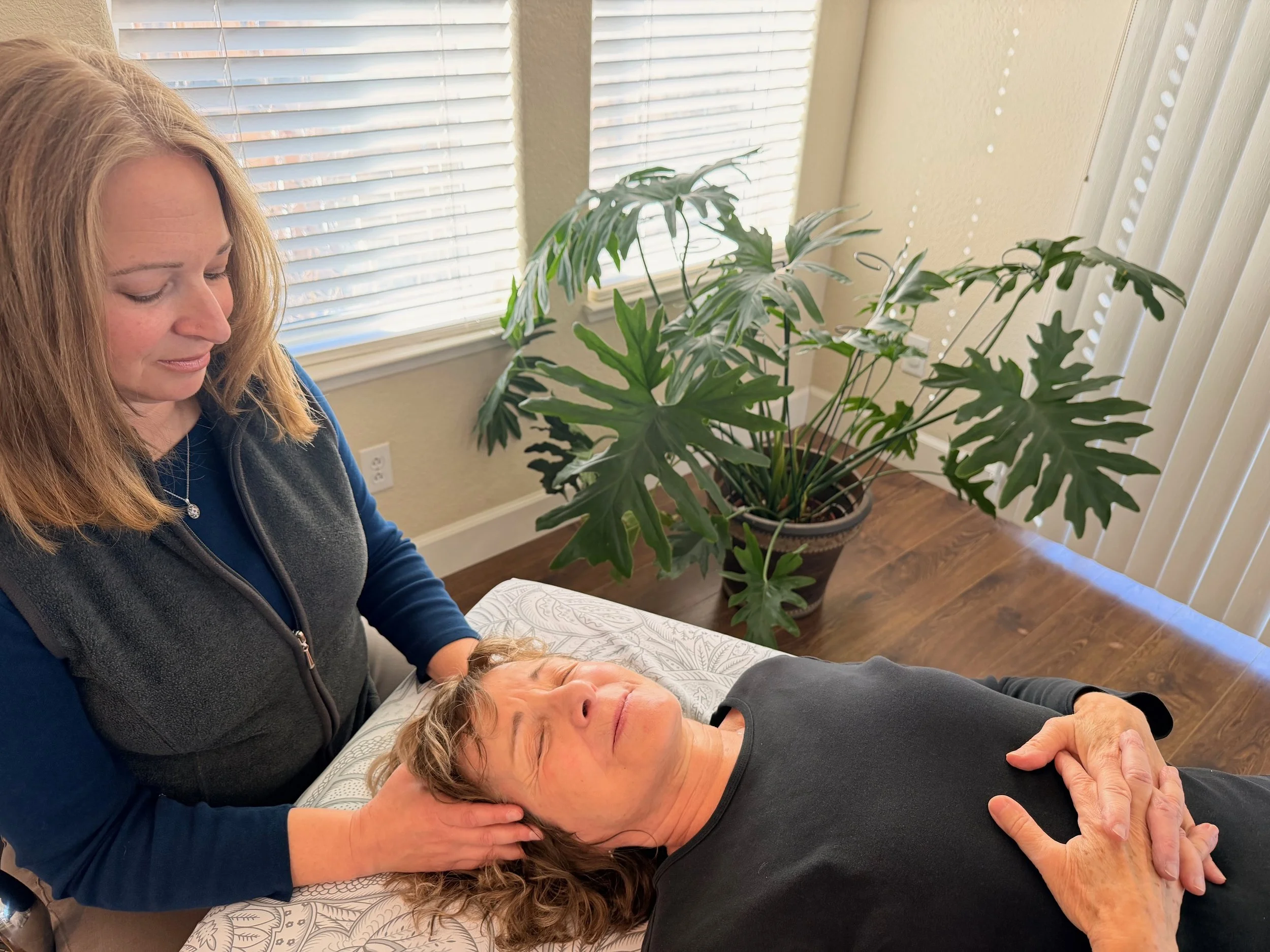 A woman gently holding a woman's head as she performs a CranioSacral Therapy treatment on a massage table in a room with large window blinds and a potted plant in the background.