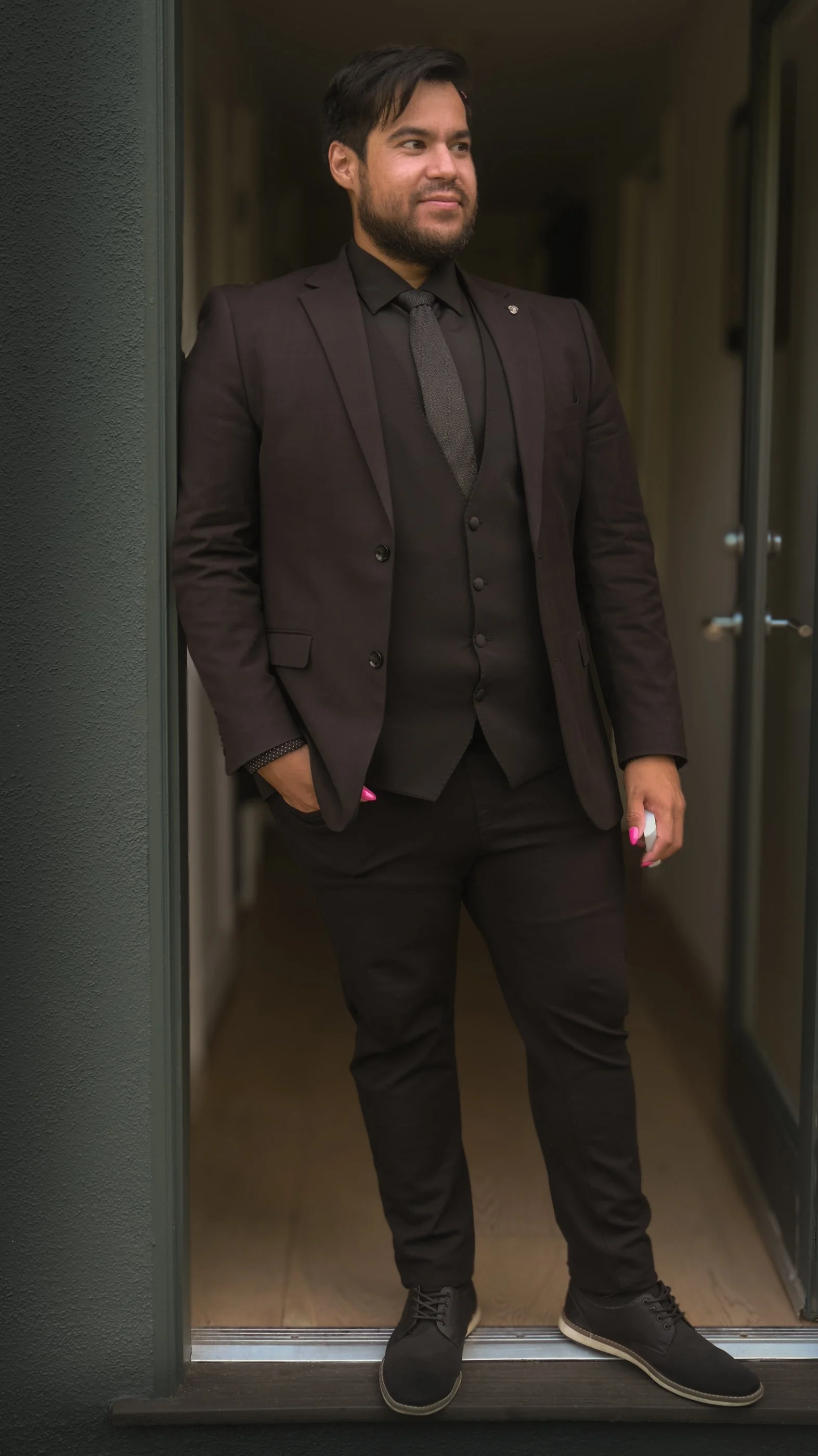 Man standing at an elevator doorway, wearing a black suit with a tie, having dark hair and a beard, with his left hand in his pocket and holding a smartphone in his right hand.