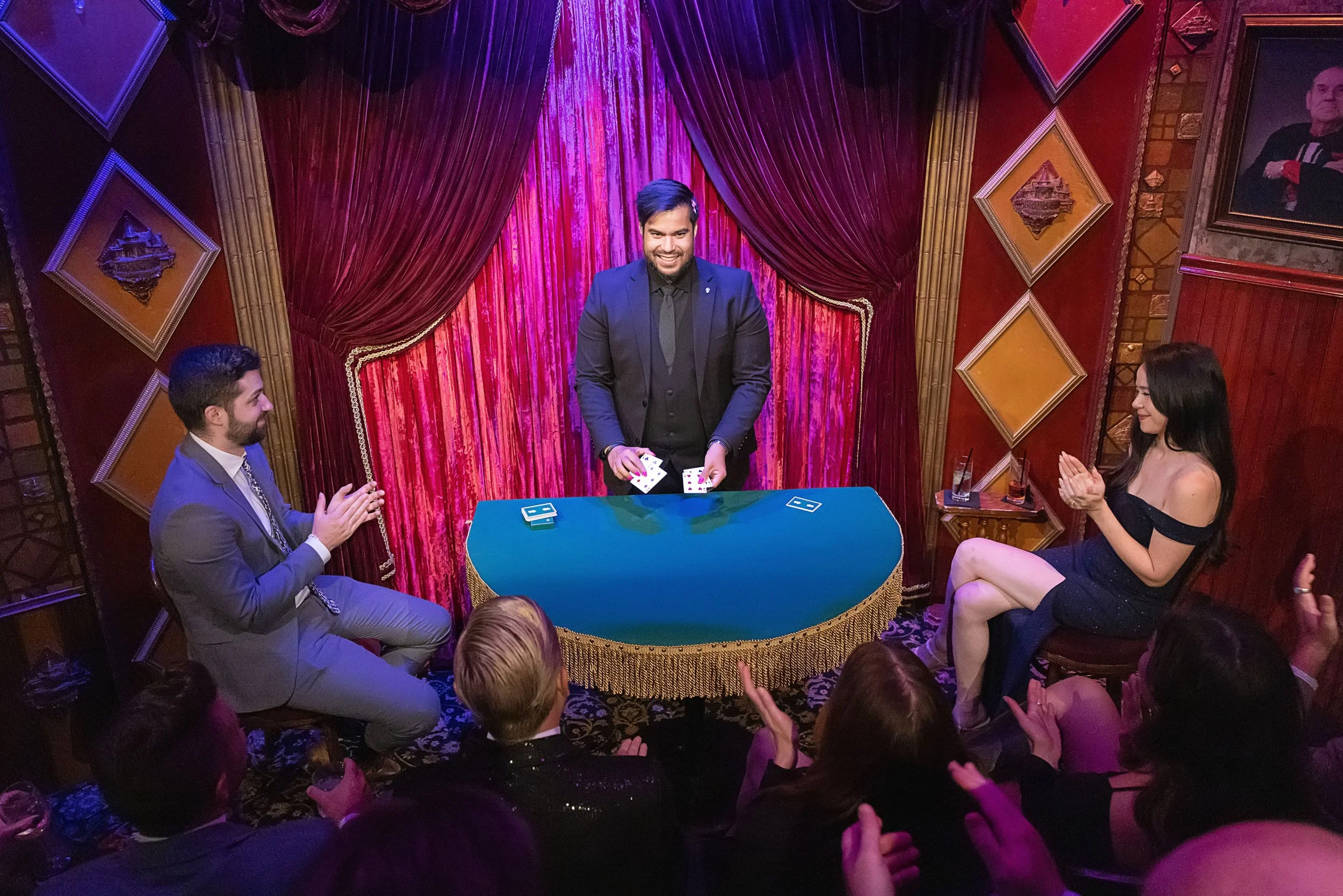 A magician performs a card trick on a stage with red drapes and gold decorative framing, while an audience applauds and watches.
