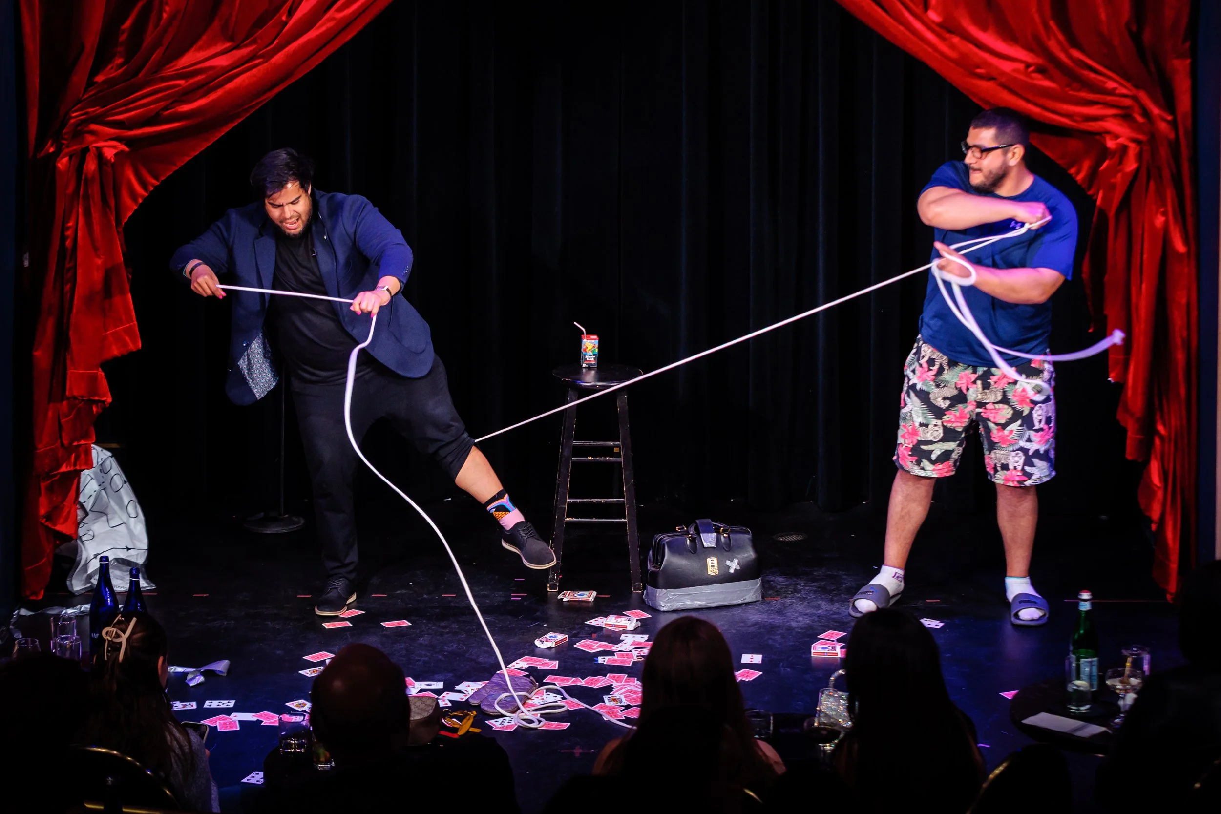 Two performers on stage engaged in a web of long strings, with scattered playing cards on the floor and an audience watching.
