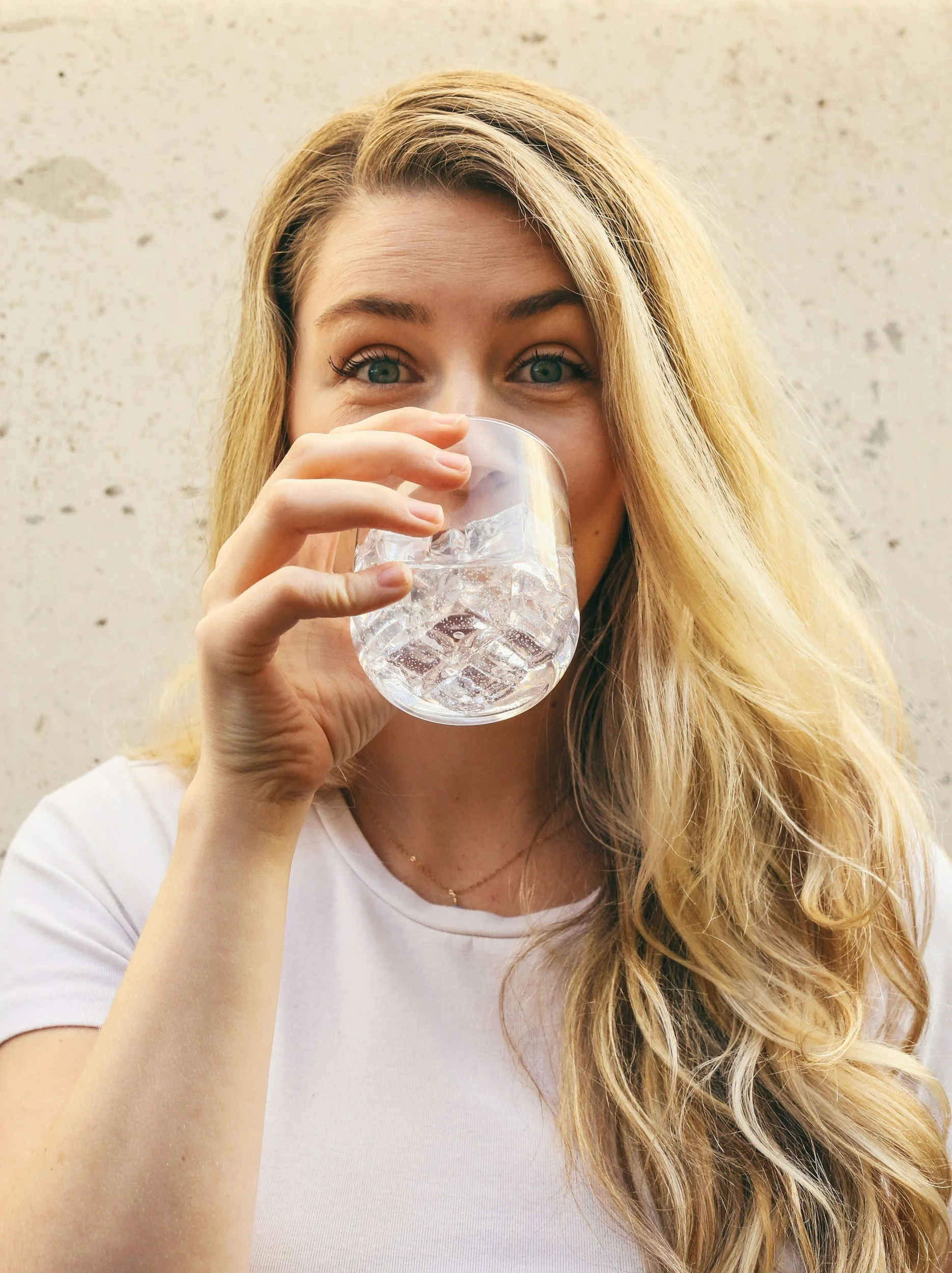Young woman with long blonde hair drinking clear water from a glass.
