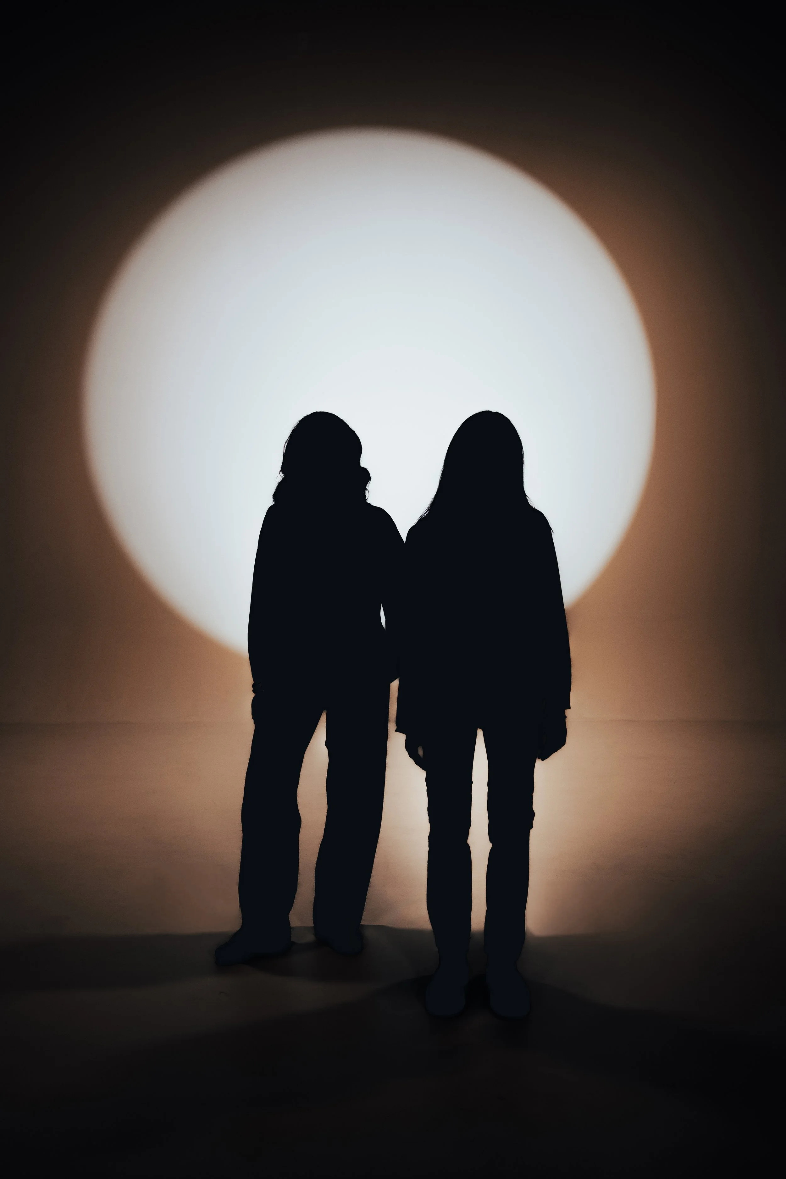 Two people in silhouette standing close together in front of a large, bright circular light on the wall, creating a shadowed effect.