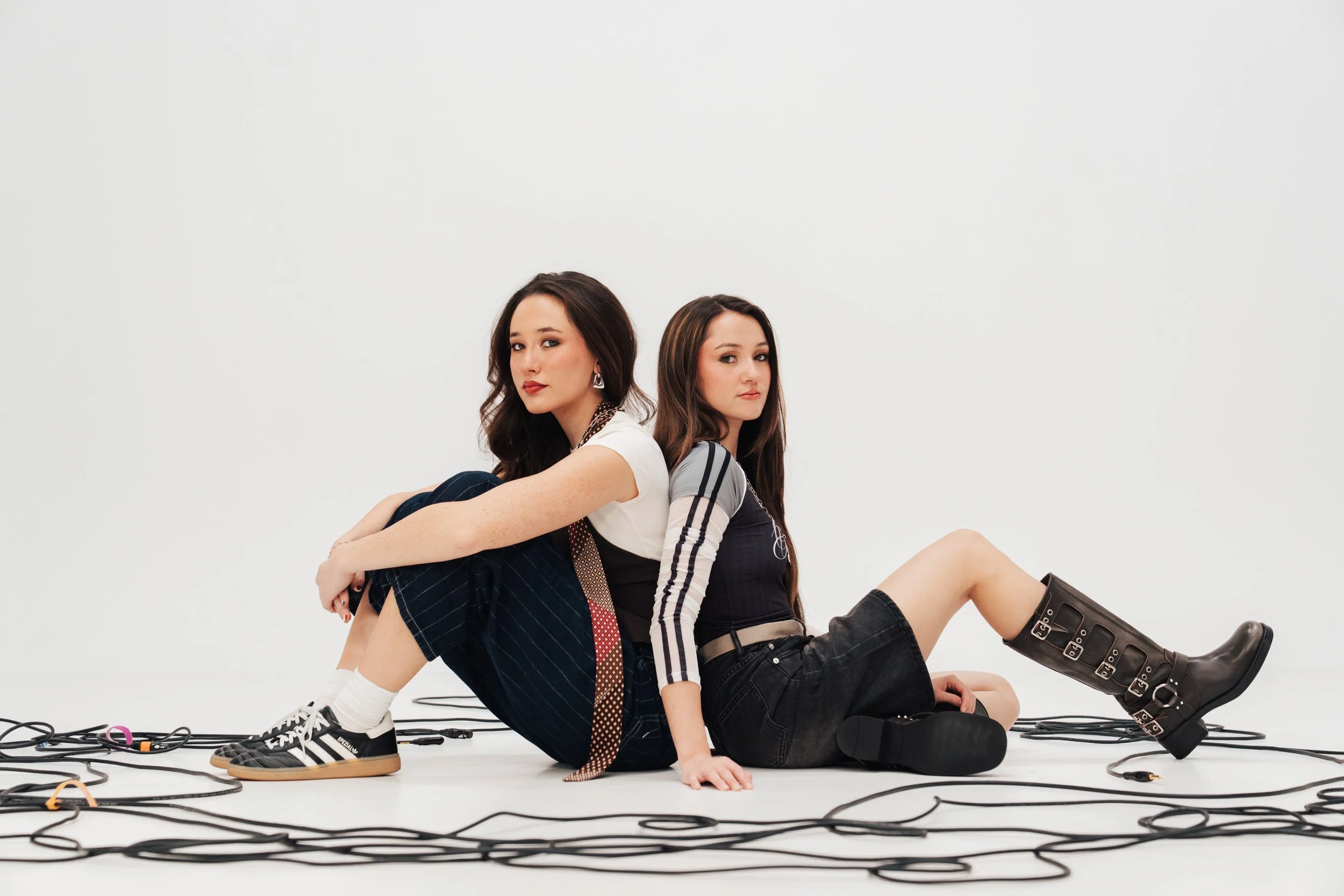 Two young women sit back-to-back on the floor surrounded by black wires against a plain white background.