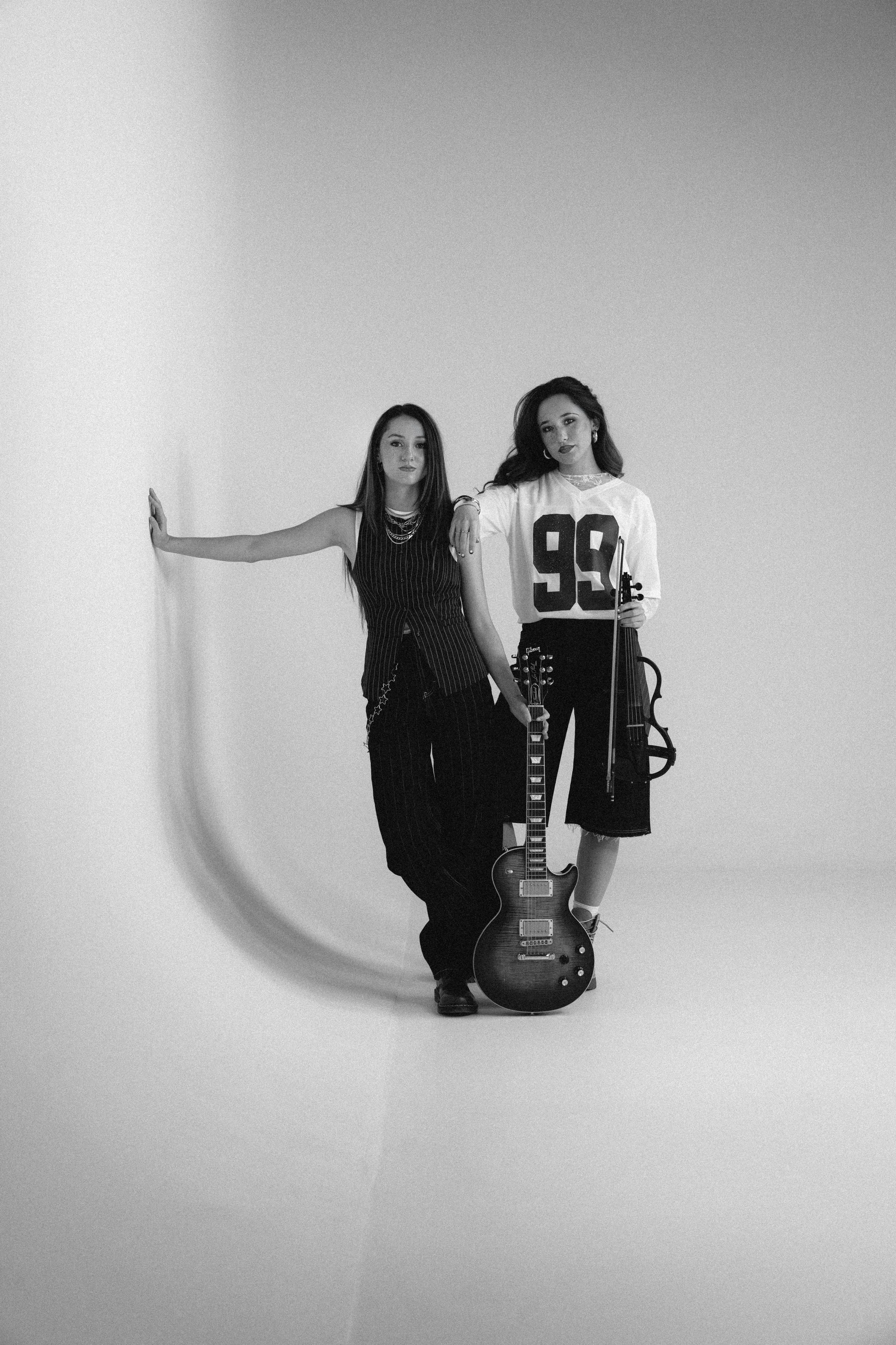 Two women standing with musical instruments in a studio. One holds a guitar, the other a violin. Both are dressed in stylish clothing, one wearing a striped dress and the other a sports jersey, with a plain backdrop.