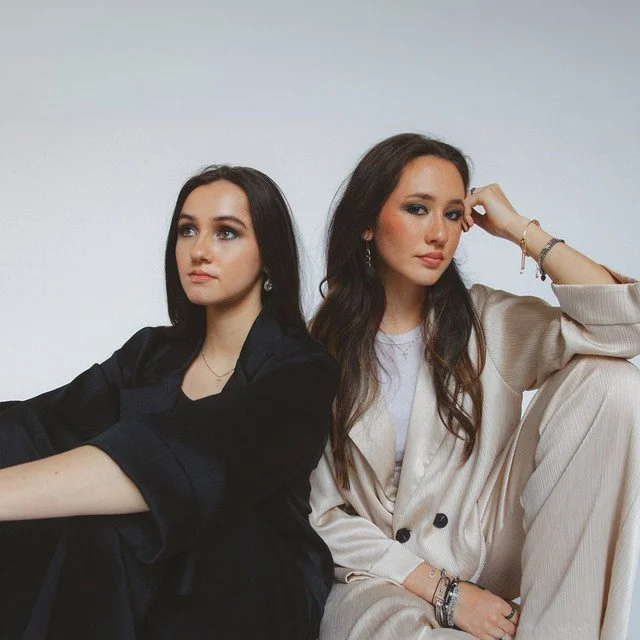 Two young women sitting side by side against a plain light-colored background, dressed in fashionable casual clothing.