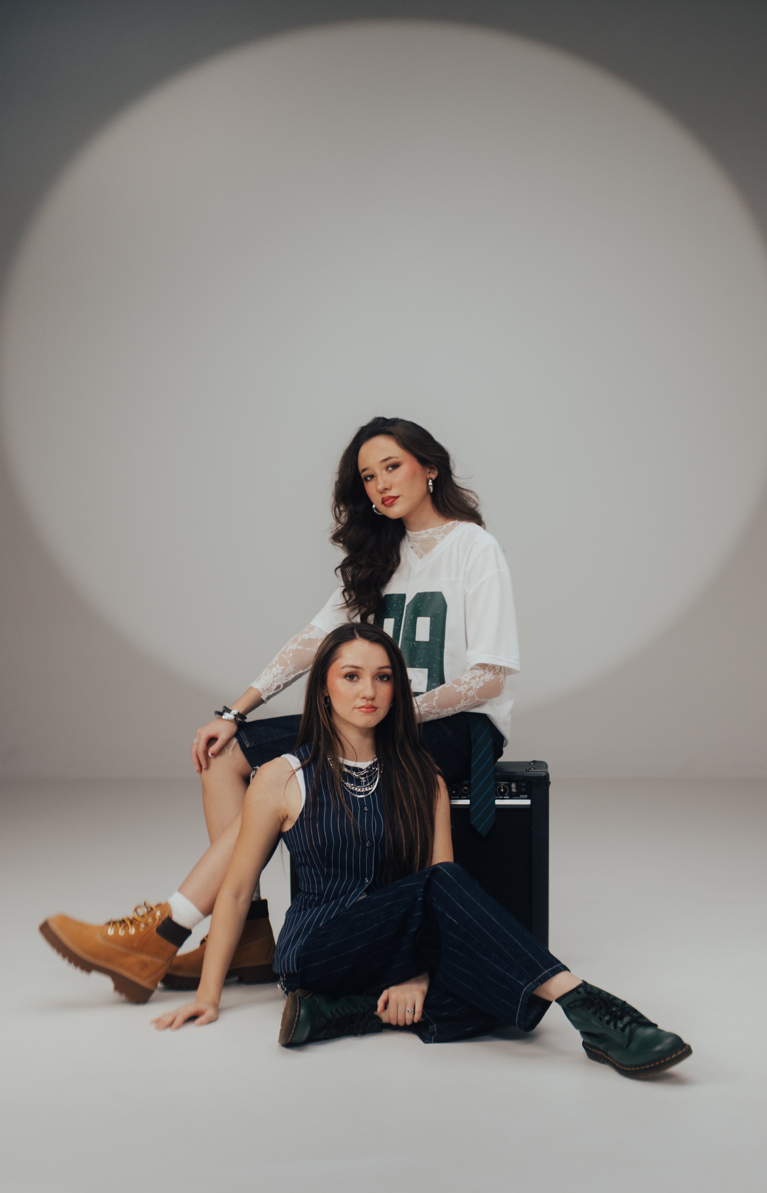 Two young women with long hair, one seated on the floor and the other sitting on a speaker, in a photography studio with a plain background and a circular spotlight. They are dressed in casual and trendy outfits, with accessories and footwear, posing