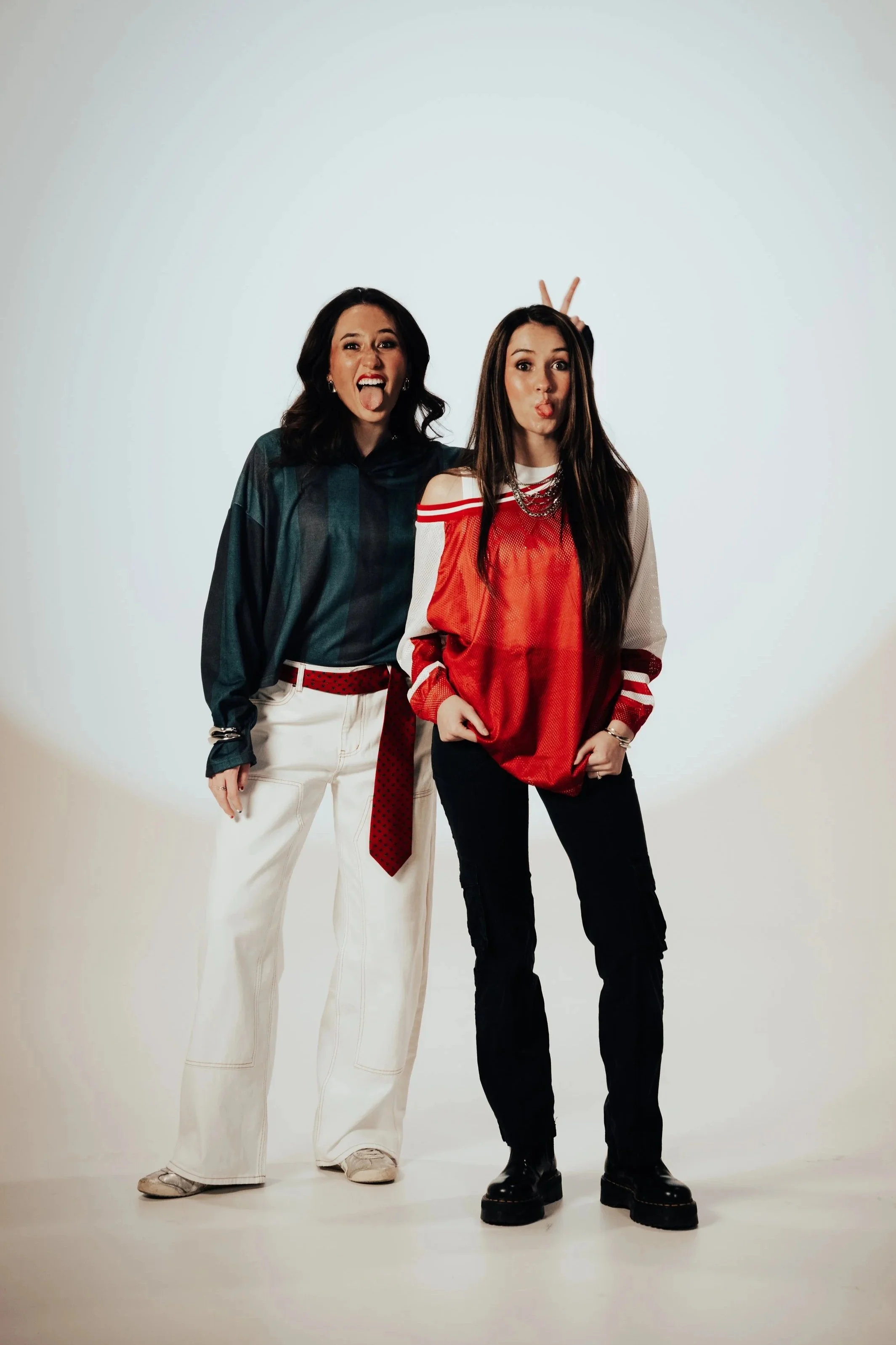 Two women posing playfully, one giving bunny ears behind the other, both making funny faces in a studio with a plain background.