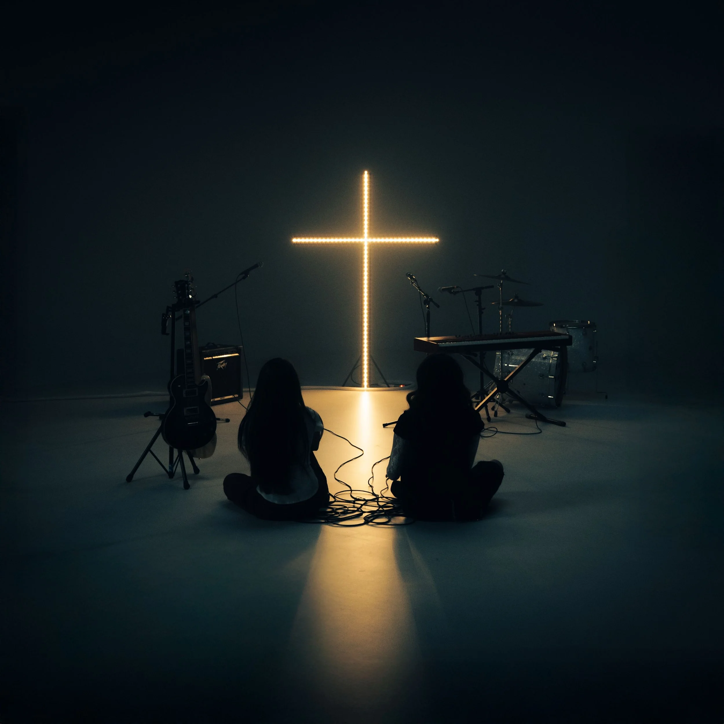 Two individuals sitting on the floor facing a lit cross-shaped light installation, with musical instruments including guitars, microphones, a keyboard, drums, and amplifiers nearby in a dark room.