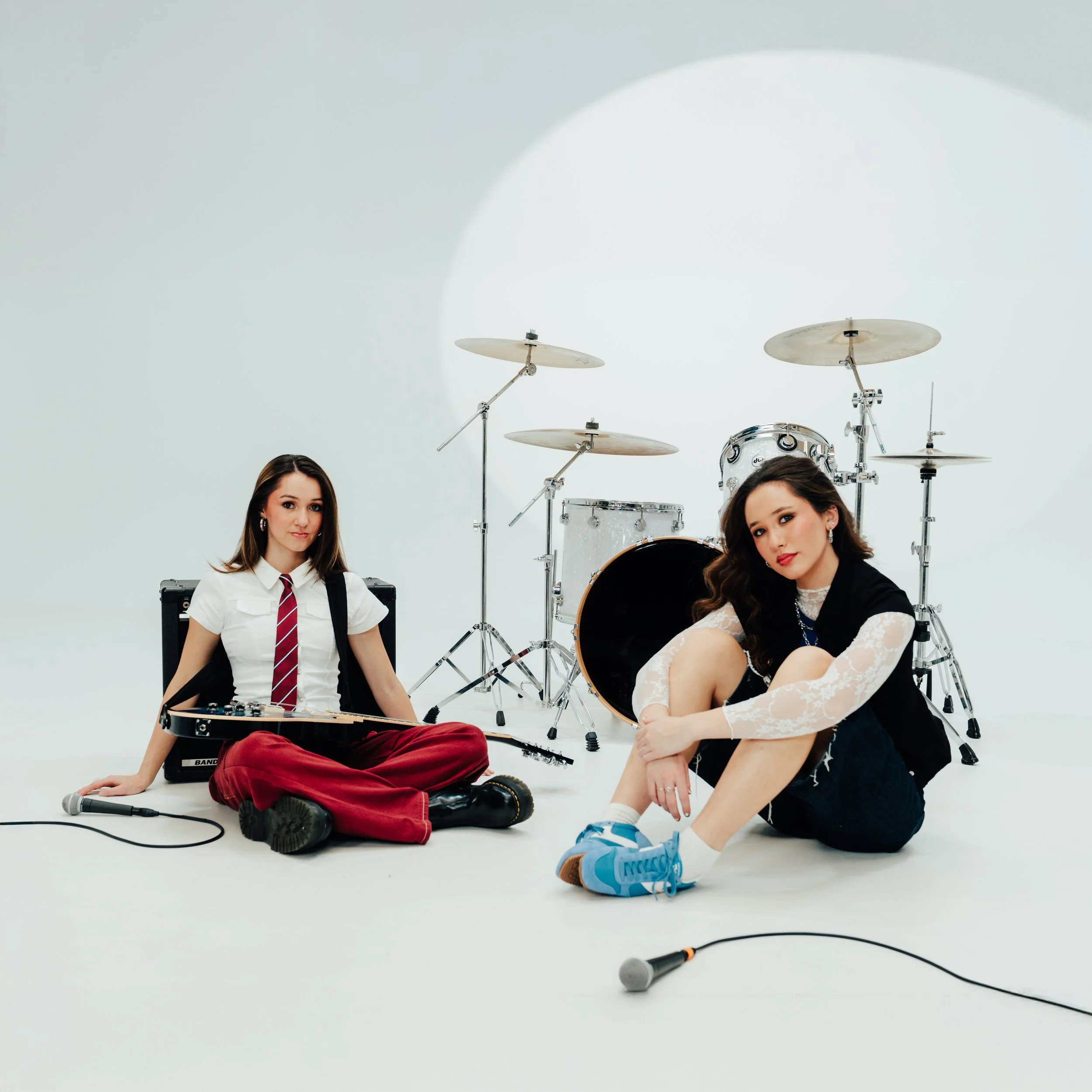 Two young women sitting on the floor in front of a drum set and musical equipment, in a white studio background.