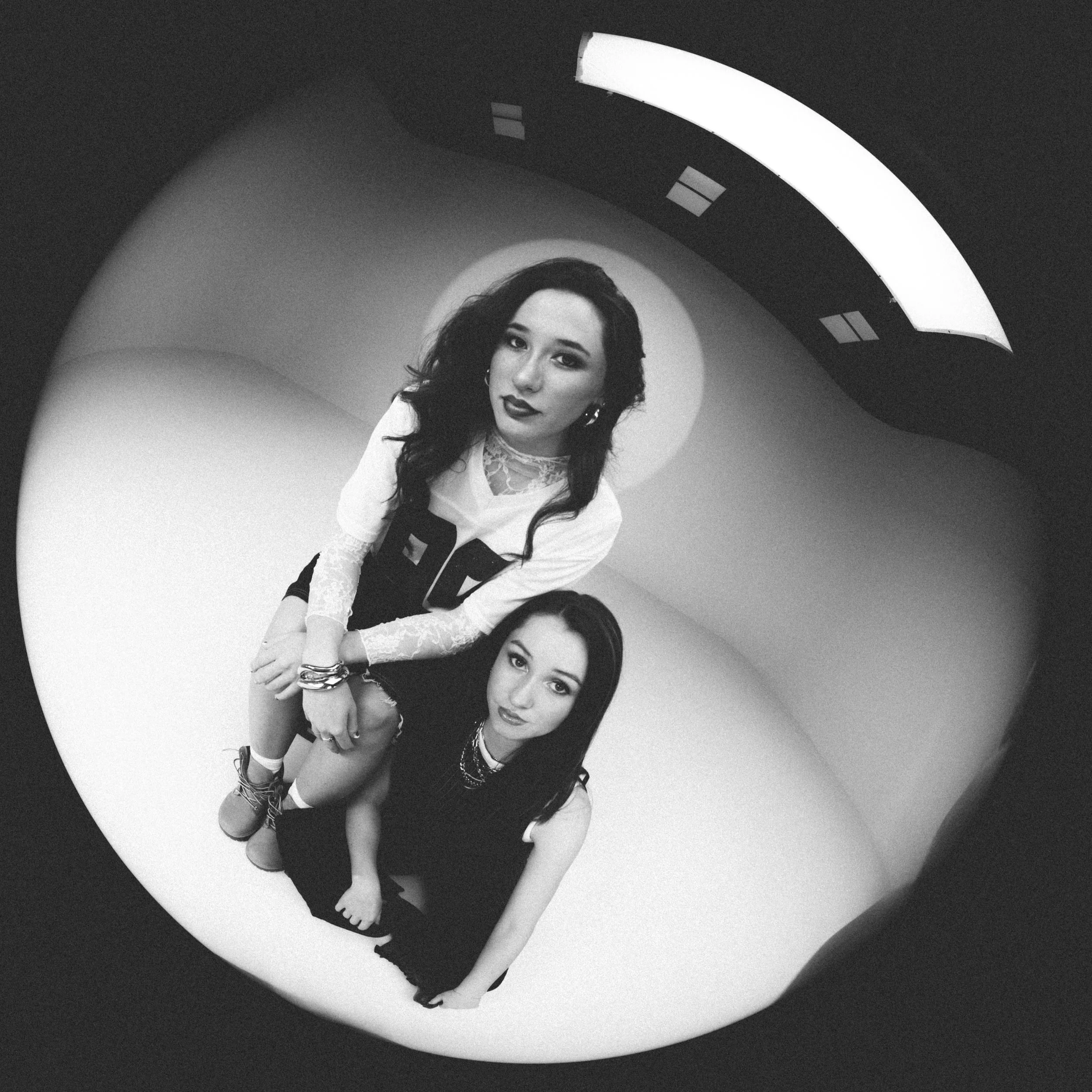 Black and white photograph of two women looking up at the camera through a fisheye lens. The woman on top has dark hair, dark lipstick, and is wearing a white top with lace sleeves and dark pants. The woman on the bottom has dark hair, wearing dark c