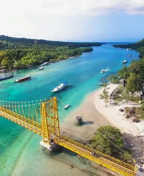 Yellow suspension bridge over a turquoise river with boats and a sandy beach, surrounded by green trees and hills under a partly cloudy sky.