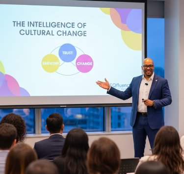 A male speaker in a blue suit giving a presentation on cultural change to an audience in a conference room, with a slide showing a diagram of trust, change, empathy, and their connections.