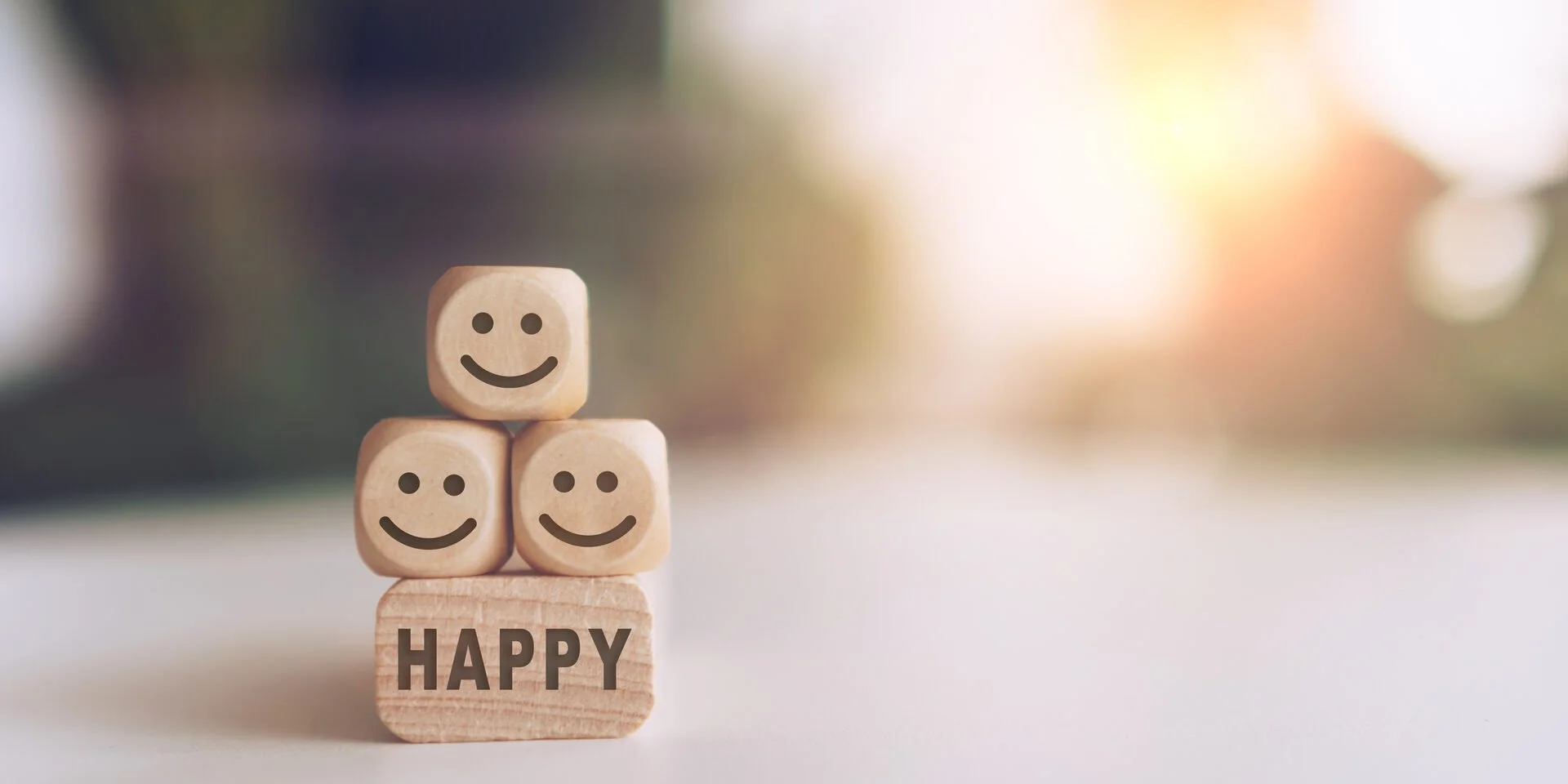 Wooden blocks with smiley faces stacked to spell out "HAPPY" on a white surface, with a blurred colorful background.