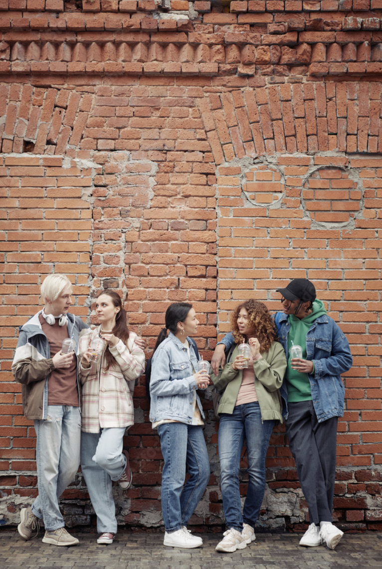 Five diverse young adults standing against a brick wall, holding drinks, and chatting casually.
