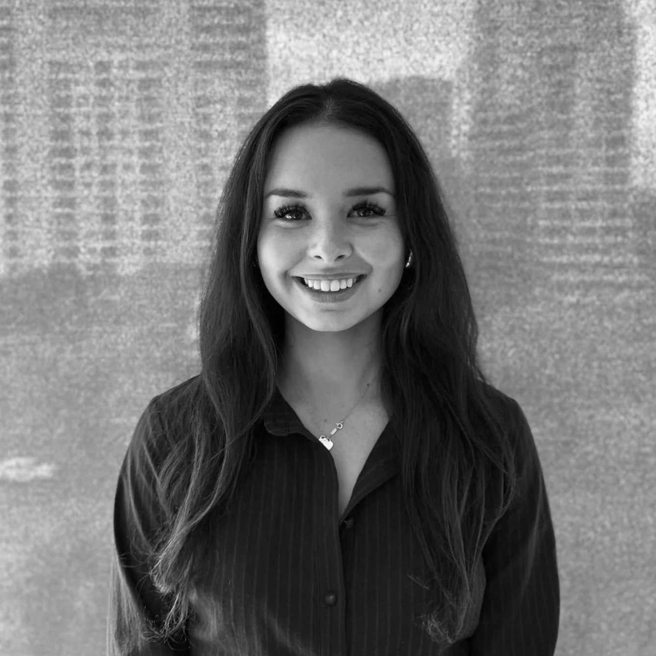 Black and white photo of a smiling woman with long dark hair, wearing a dark collared shirt, standing in front of a textured wall.