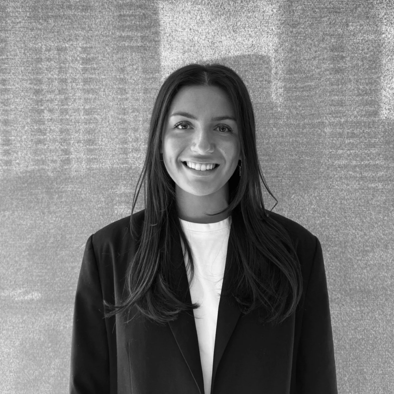 Black and white photo of a young woman with long dark hair smiling, wearing a dark blazer over a white shirt, standing in front of a textured wall.