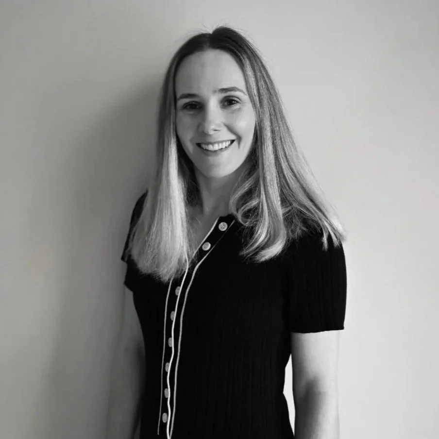 A smiling woman with long blonde hair, wearing a black short-sleeved top with a row of buttons, standing against a plain wall.