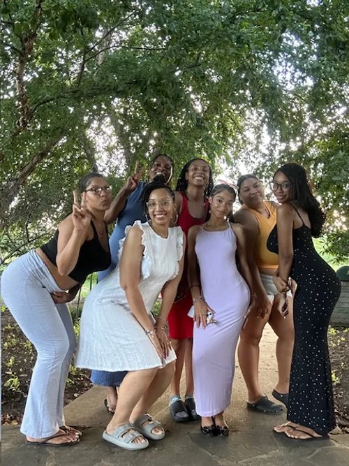 Six women posing under a large leafy tree in a park, smiling and making peace signs.