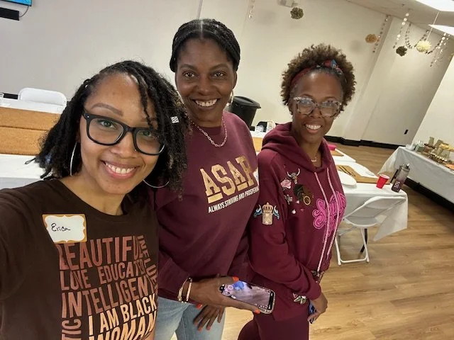 Three women smiling and standing together indoors, with tables and chairs in the background.