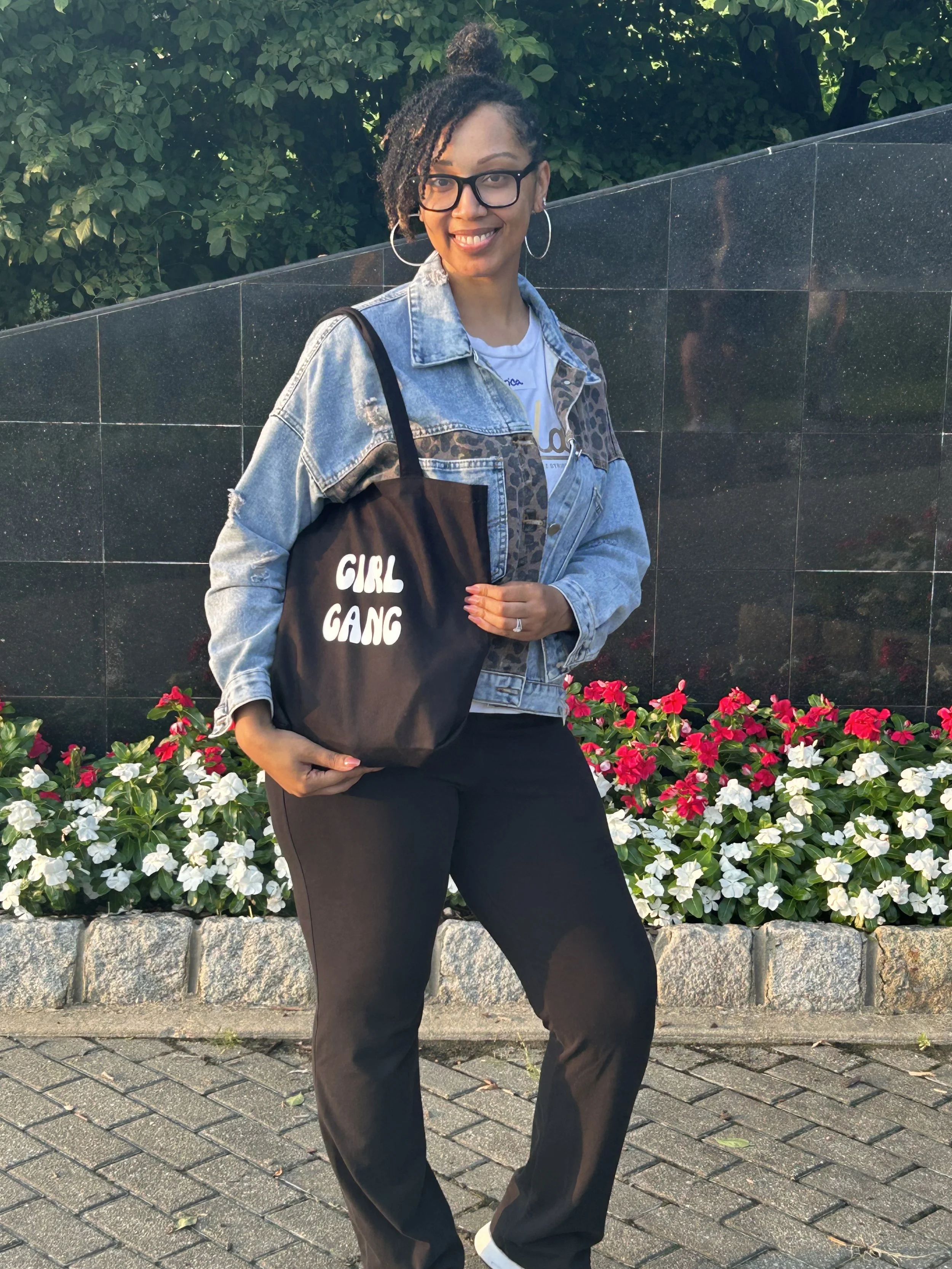 A woman with glasses and hoop earrings standing outdoors, holding a black tote bag with white text that reads 'GIRL GANG', smiling, in front of a flower bed with pink and white flowers and a dark tiled wall.